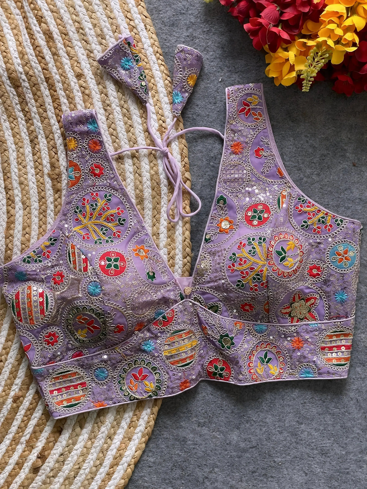 Traditional lavender blouse featuring vibrant multicolor thread embroidery and mirror work in circular floral and geometric patterns, with sequin accents and a deep V-neck tie-back detail, displayed flat on a textured mat with artificial red and yellow flowers in the corner.