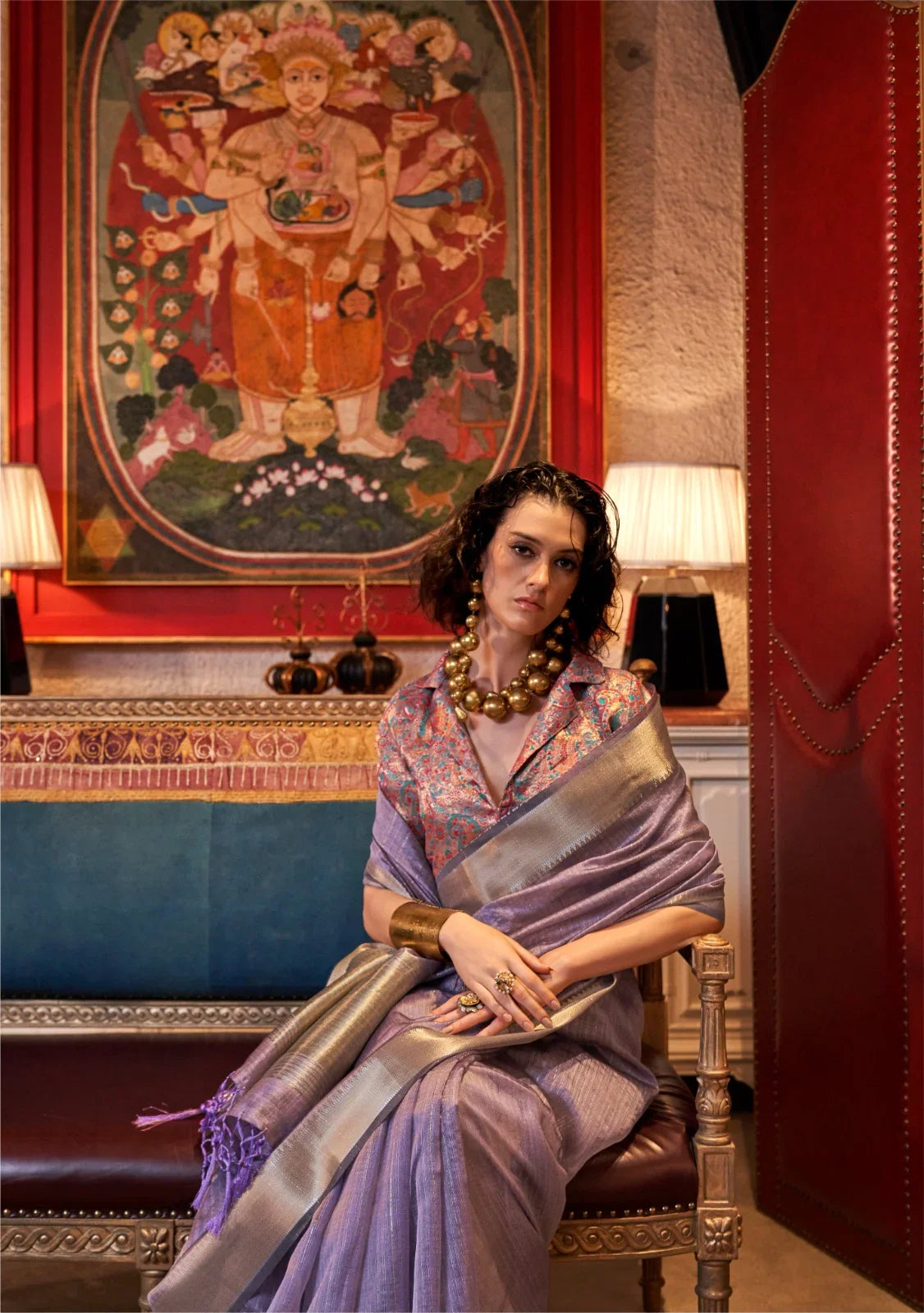 Elegant woman in lavender saree with intricate zari work and embroidered blouse, seated in a luxurious setting showcasing ethnic fashion