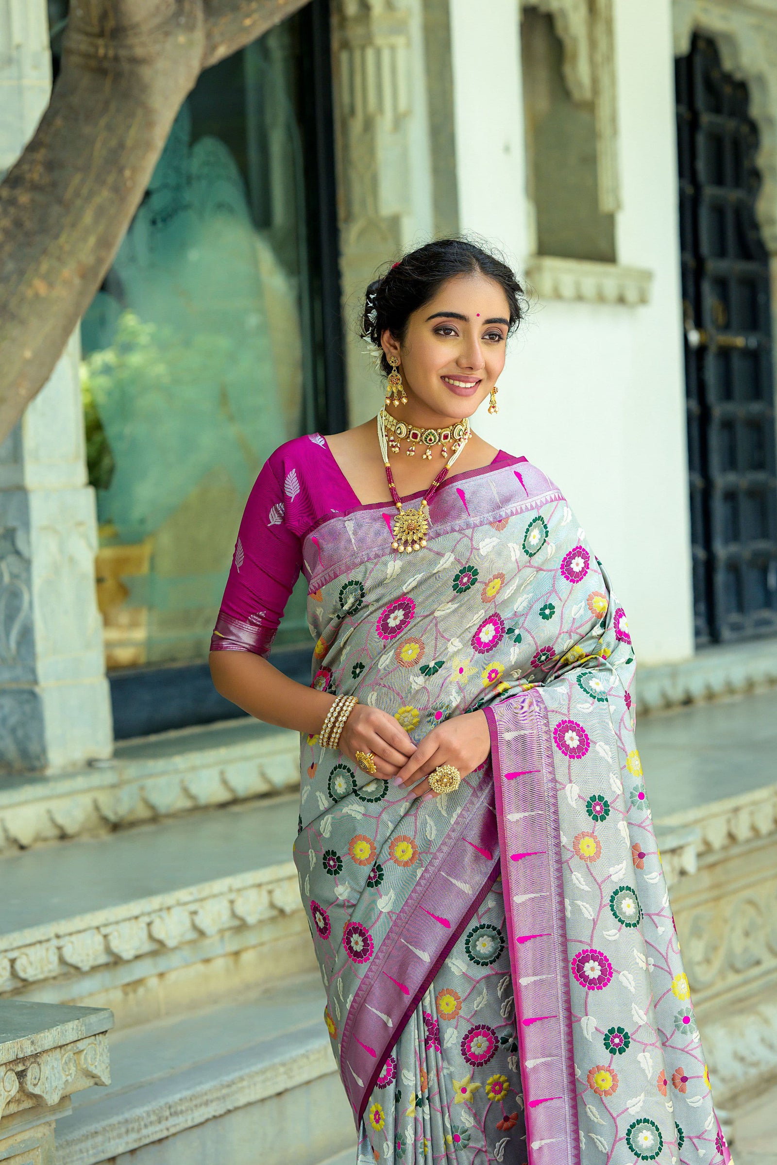 Smiling woman wearing light gray zari work saree with pink blouse and traditional jewelry in ethnic setting