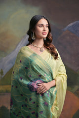 A woman models a soft Pista Green Muga Silk Saree with subtle Resham weaving. The pallu dramatically transitions to a deep teal color, featuring intricate purple and green floral/peacock Resham threadwork. She wears a teal blouse and an oxidized silver necklace.
