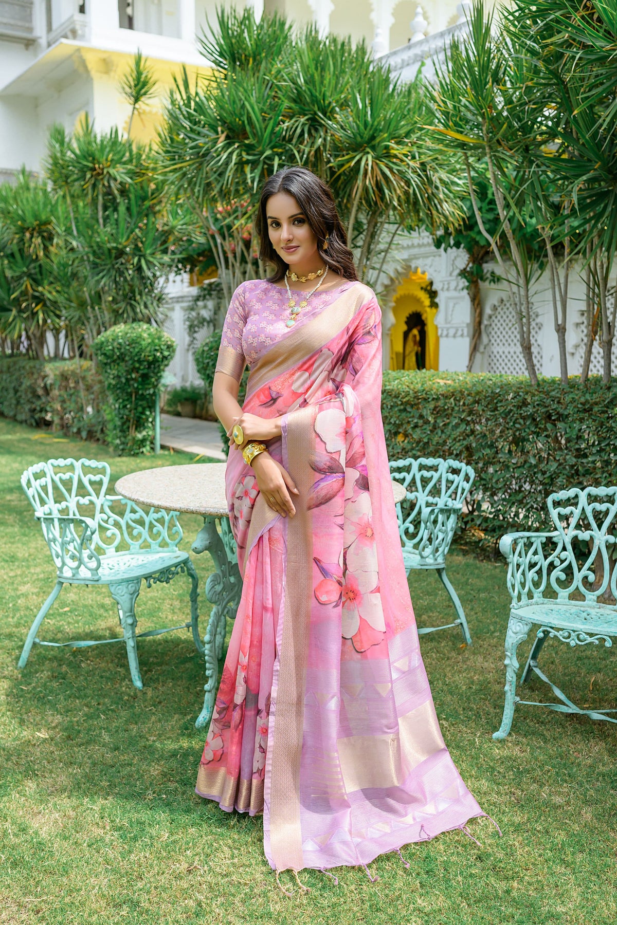 Woman wearing light pink zari work saree with floral patterns and golden border in a royal garden setting