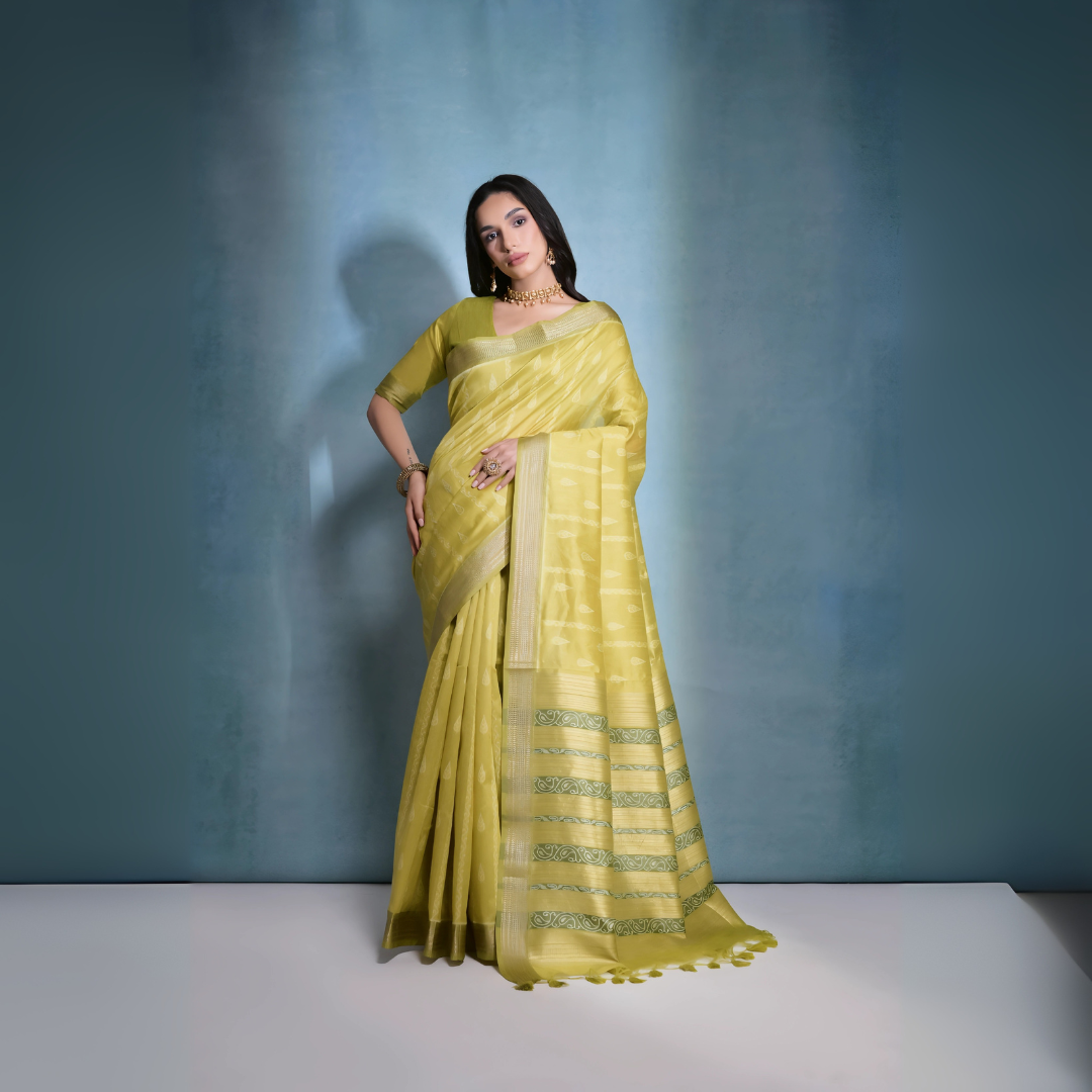 A woman models an elegant Light Yellow/Lime Green Soft Raw Silk Saree with a wide metallic gold Zari border. The pallu features distinct horizontal bands of green and gold Ikat-style geometric weaving. She wears a matching light green blouse and accessorizes with a gold choker necklace and large gold earrings, posing indoors.