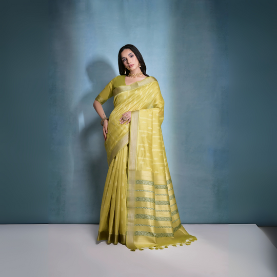 A woman models an elegant Light Yellow/Lime Green Soft Raw Silk Saree with a wide metallic gold Zari border. The pallu features distinct horizontal bands of green and gold Ikat-style geometric weaving. She wears a matching light green blouse and accessorizes with a gold choker necklace and large gold earrings, posing indoors.