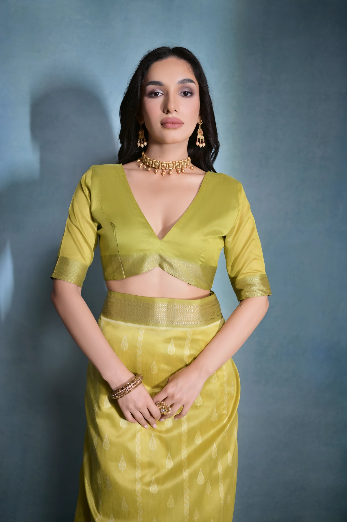 A woman models an elegant Light Yellow/Lime Green Soft Raw Silk Saree with a wide metallic gold Zari border. The pallu features distinct horizontal bands of green and gold Ikat-style geometric weaving. She wears a matching light green blouse and accessorizes with a gold choker necklace and large gold earrings, posing indoors.