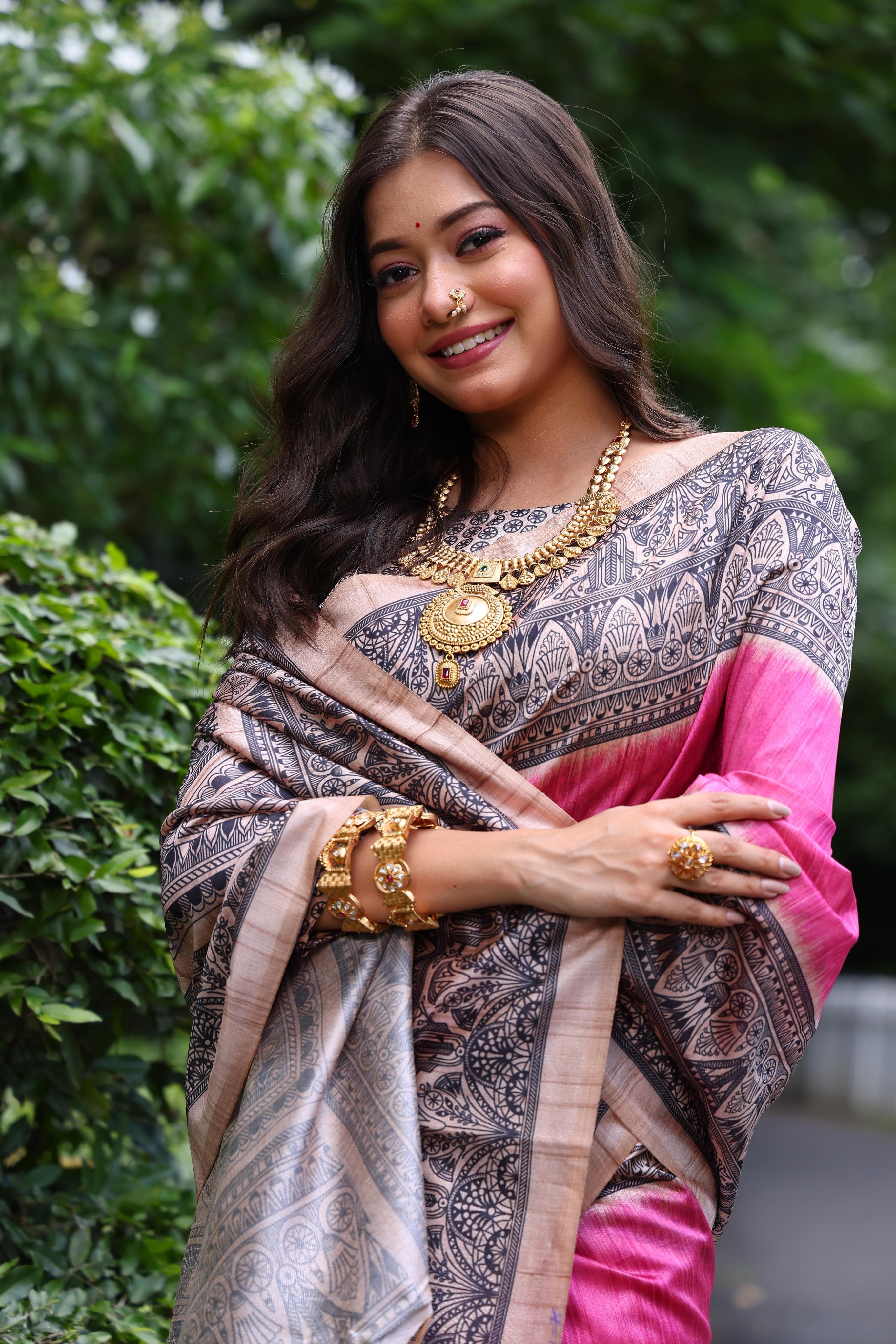 Woman wearing traditional dark pink Madhubani print saree with intricate designs and ethnic jewelry in a green outdoor setting