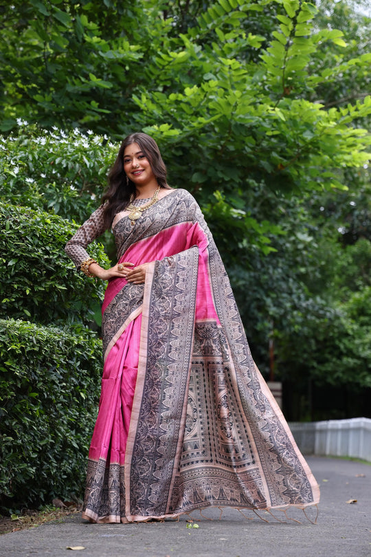 Woman wearing traditional orange Madhubani print saree with intricate designs and ethnic jewelry in a green outdoor setting
