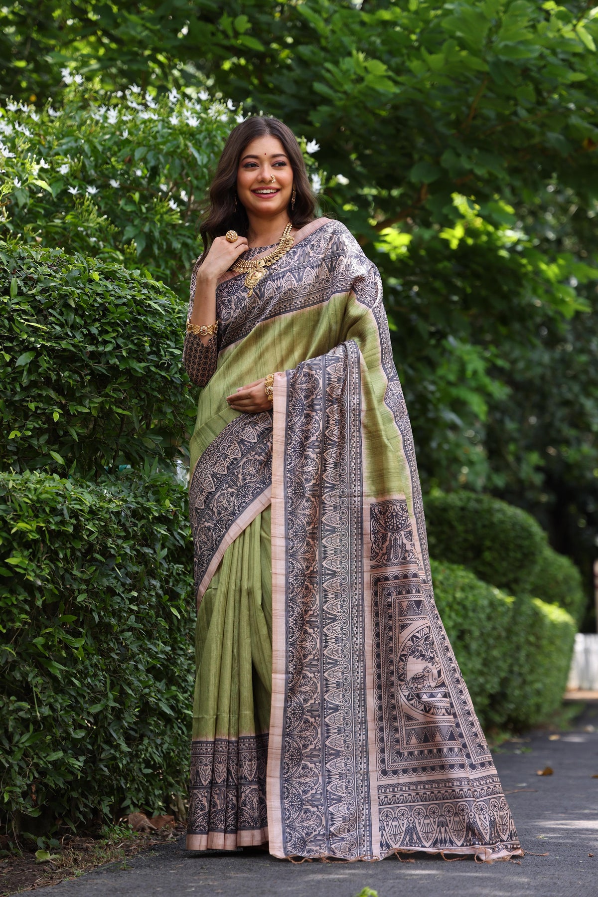 Woman wearing traditional olivine Madhubani print saree with intricate designs and ethnic jewelry in a green outdoor setting