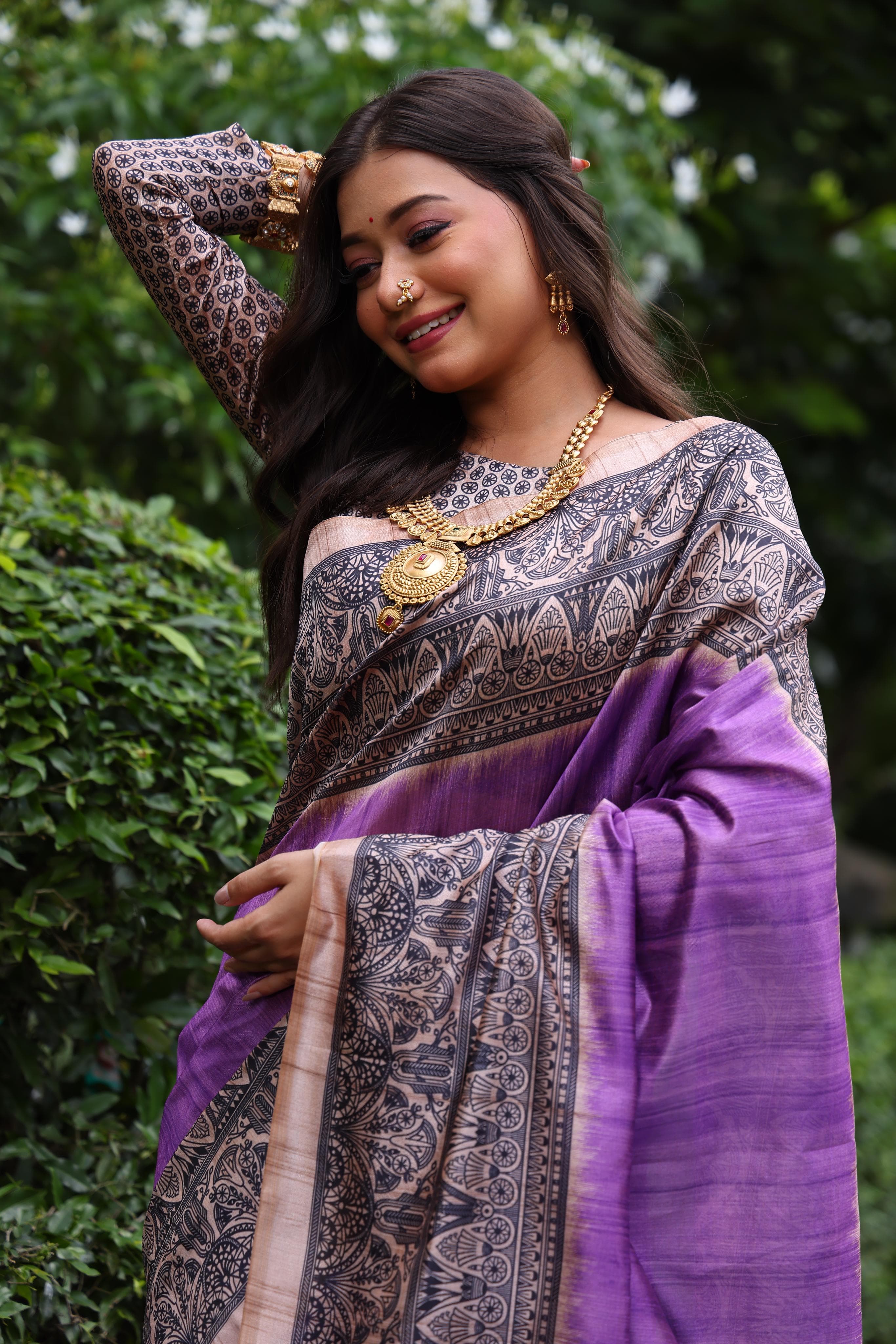 Woman wearing traditional purple Madhubani print saree with intricate designs and ethnic jewelry in a green outdoor setting
