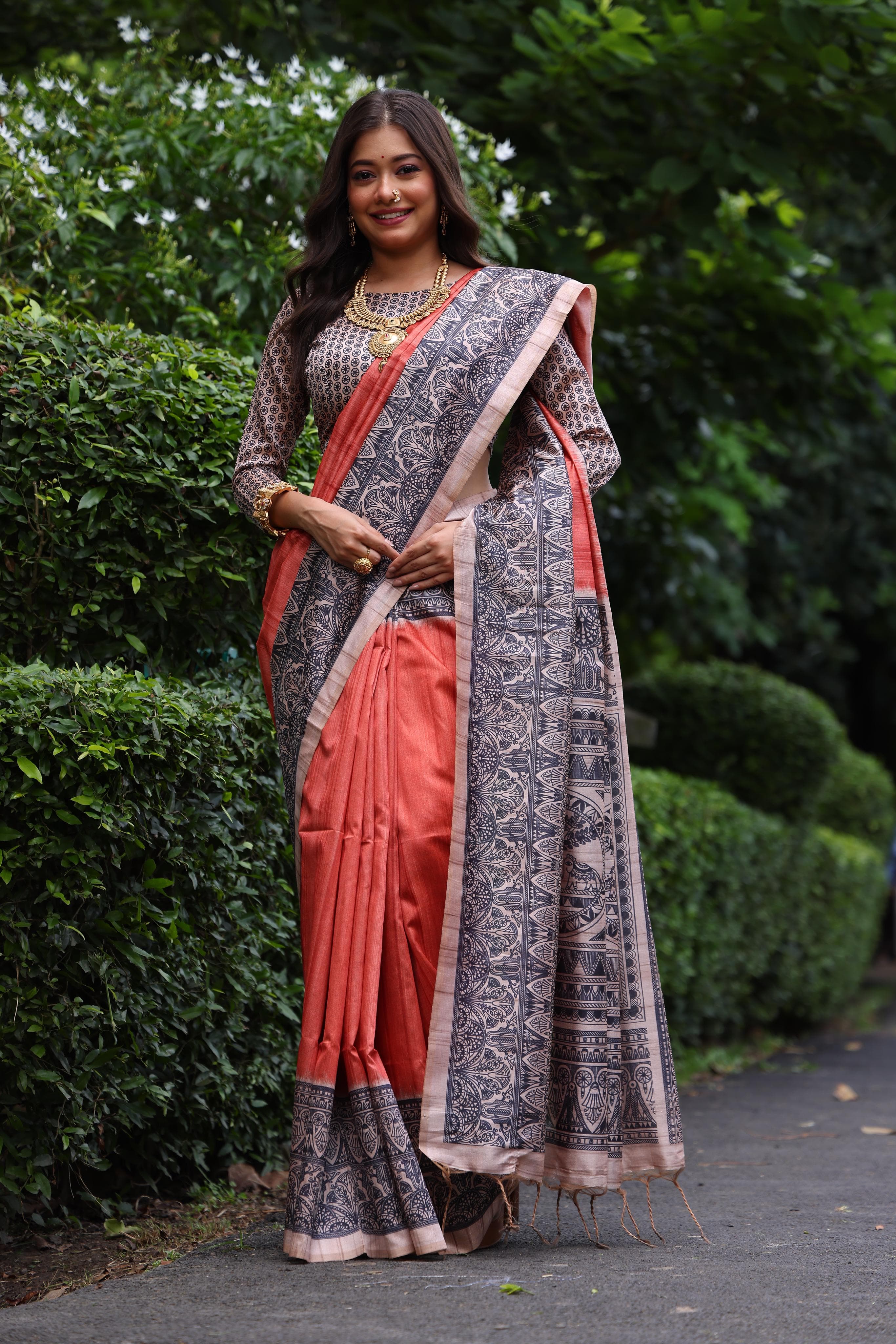 Woman wearing traditional orange Madhubani print saree with intricate designs and ethnic jewelry in a green outdoor setting