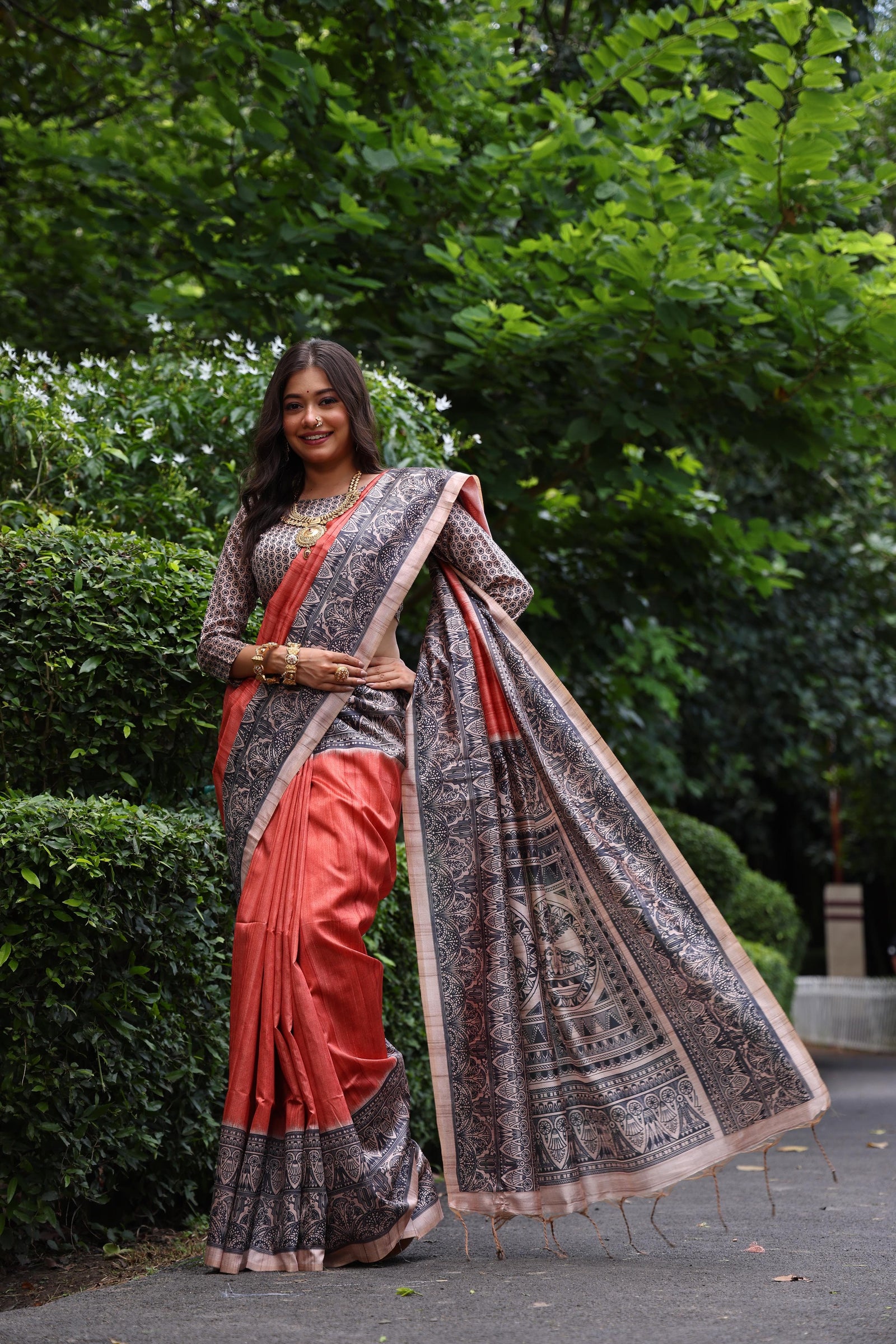 Woman wearing traditional orange Madhubani print saree with intricate designs and ethnic jewelry in a green outdoor setting
