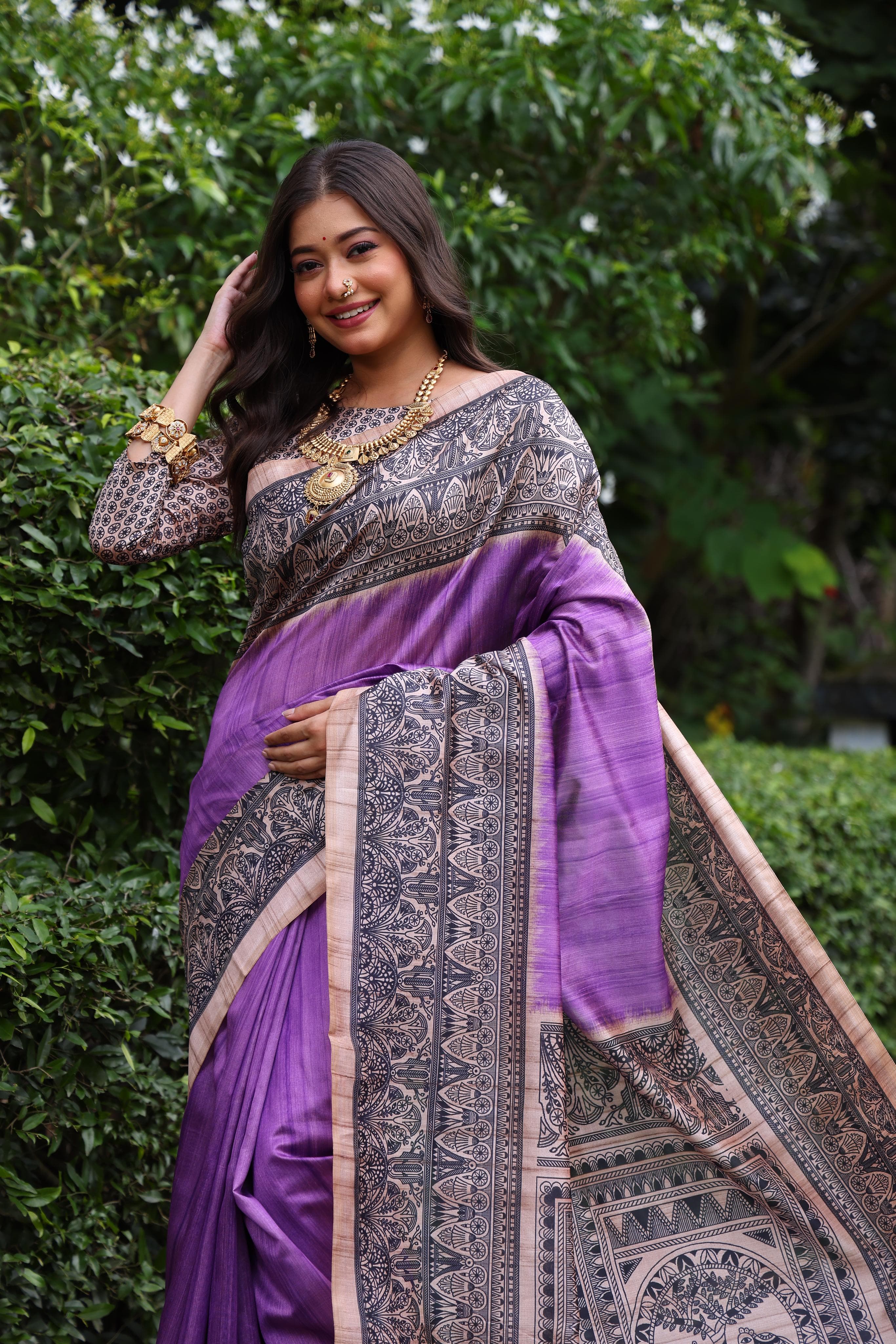 Woman wearing traditional purple Madhubani print saree with intricate designs and ethnic jewelry in a green outdoor setting