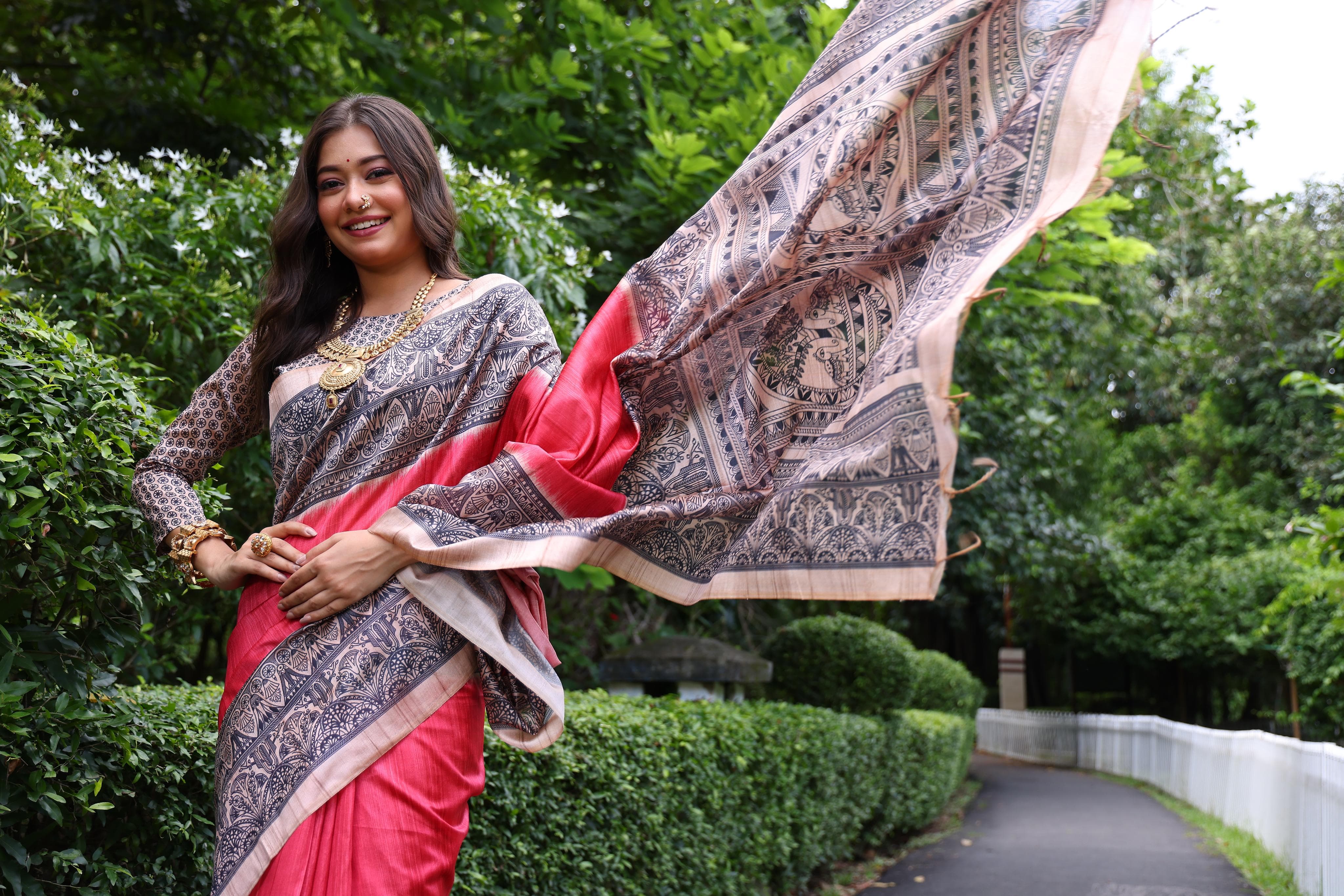Woman wearing traditional red Madhubani print saree with intricate designs and ethnic jewelry in a green outdoor setting