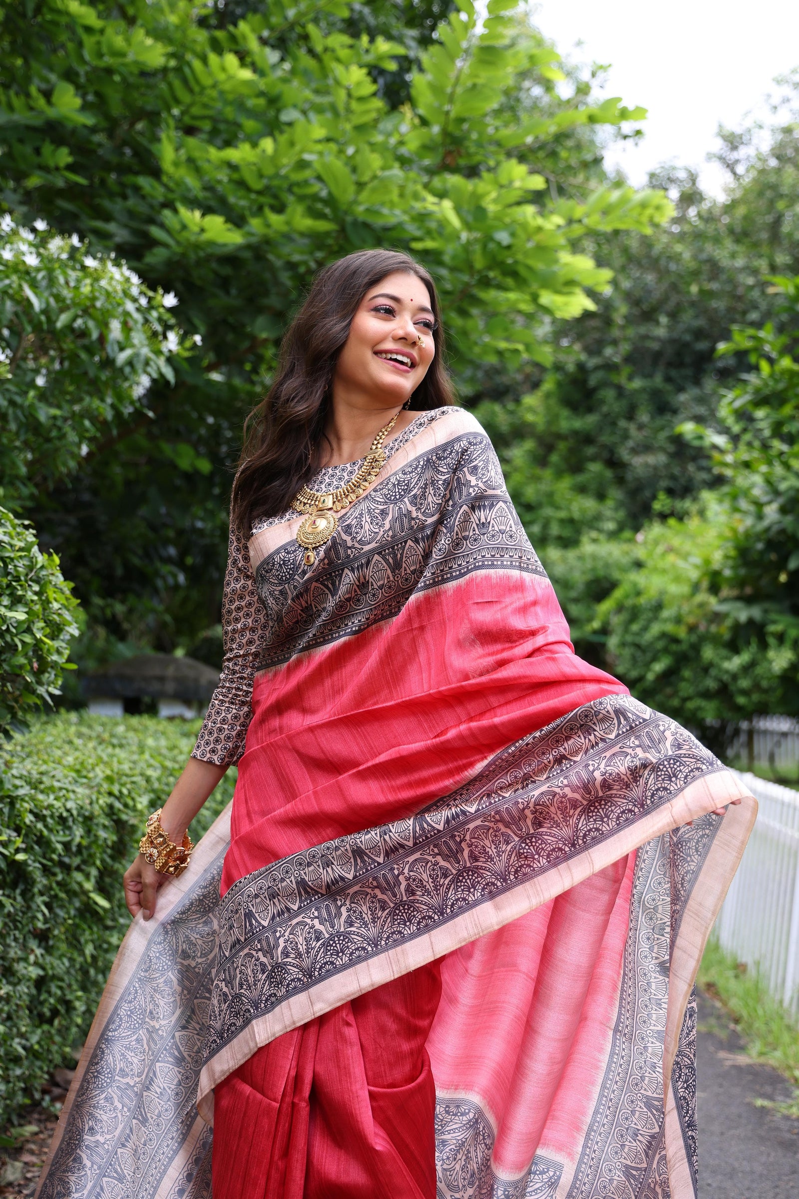 Woman wearing traditional red Madhubani print saree with intricate designs and ethnic jewelry in a green outdoor setting
