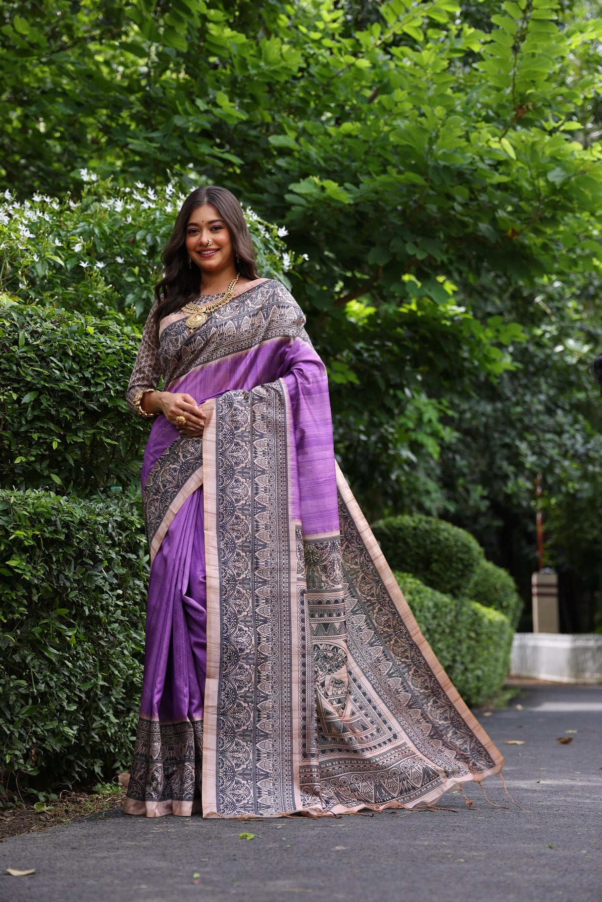 Woman wearing traditional purple Madhubani print saree with intricate designs and ethnic jewelry in a green outdoor setting