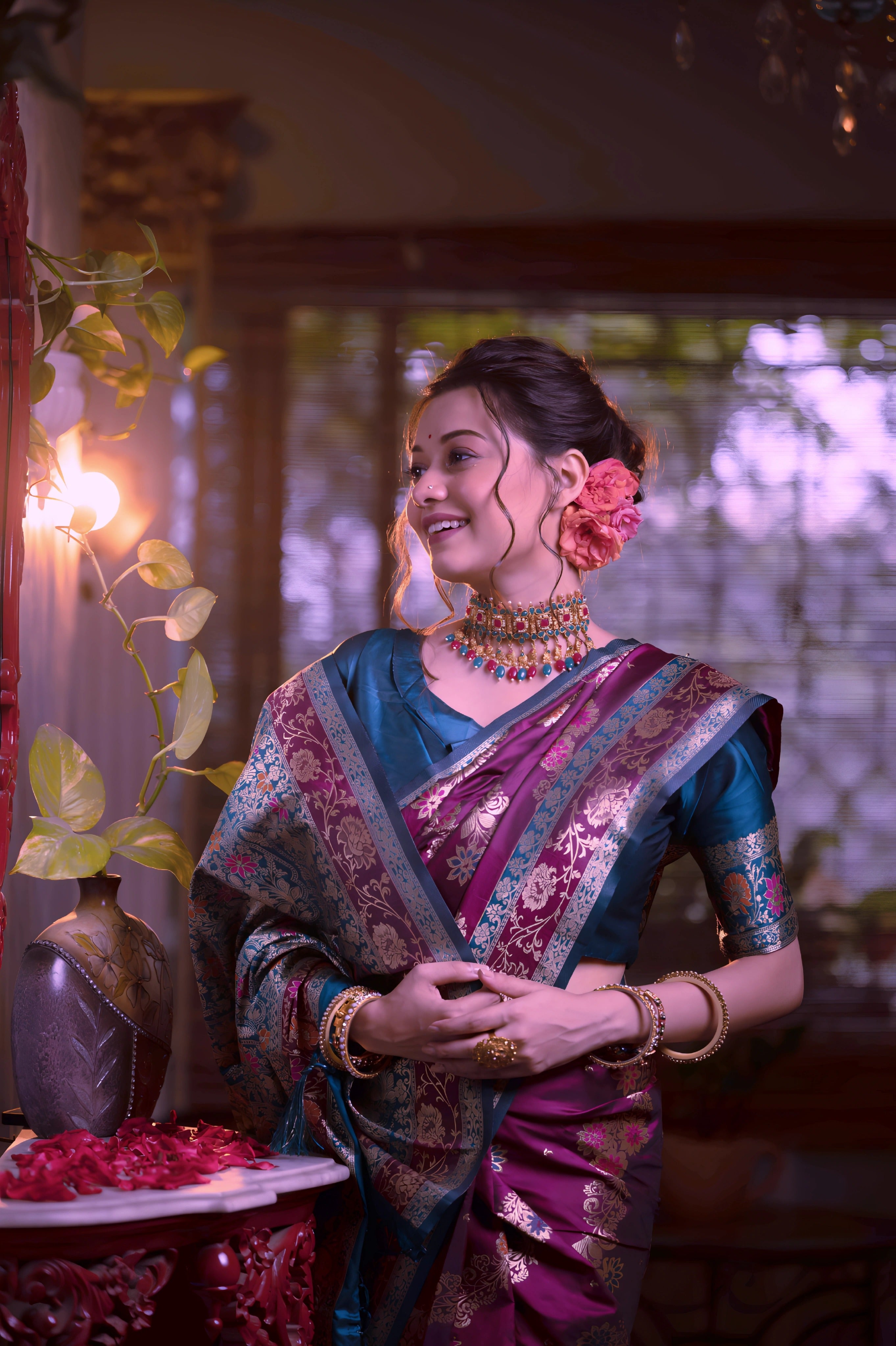 A woman models a luxurious Wine Red/Maroon Soft Banarasi Silk Saree. The saree features gold Zari weaving and a wide border with prominent Teal/Aqua Blue contrast accents. She wears a matching teal short-sleeve blouse and a gold Kundan choker necklace, posing in a dramatically lit antique room.
