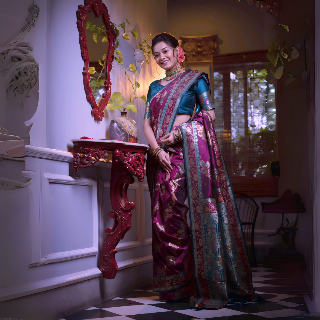 A woman models a luxurious Wine Red/Maroon Soft Banarasi Silk Saree. The saree features gold Zari weaving and a wide border with prominent Teal/Aqua Blue contrast accents. She wears a matching teal short-sleeve blouse and a gold Kundan choker necklace, posing in a dramatically lit antique room.