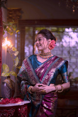 A woman models a luxurious Wine Red/Maroon Soft Banarasi Silk Saree. The saree features gold Zari weaving and a wide border with prominent Teal/Aqua Blue contrast accents. She wears a matching teal short-sleeve blouse and a gold Kundan choker necklace, posing in a dramatically lit antique room.
