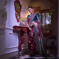 A woman models a luxurious Wine Red/Maroon Soft Banarasi Silk Saree. The saree features gold Zari weaving and a wide border with prominent Teal/Aqua Blue contrast accents. She wears a matching teal short-sleeve blouse and a gold Kundan choker necklace, posing in a dramatically lit antique room.