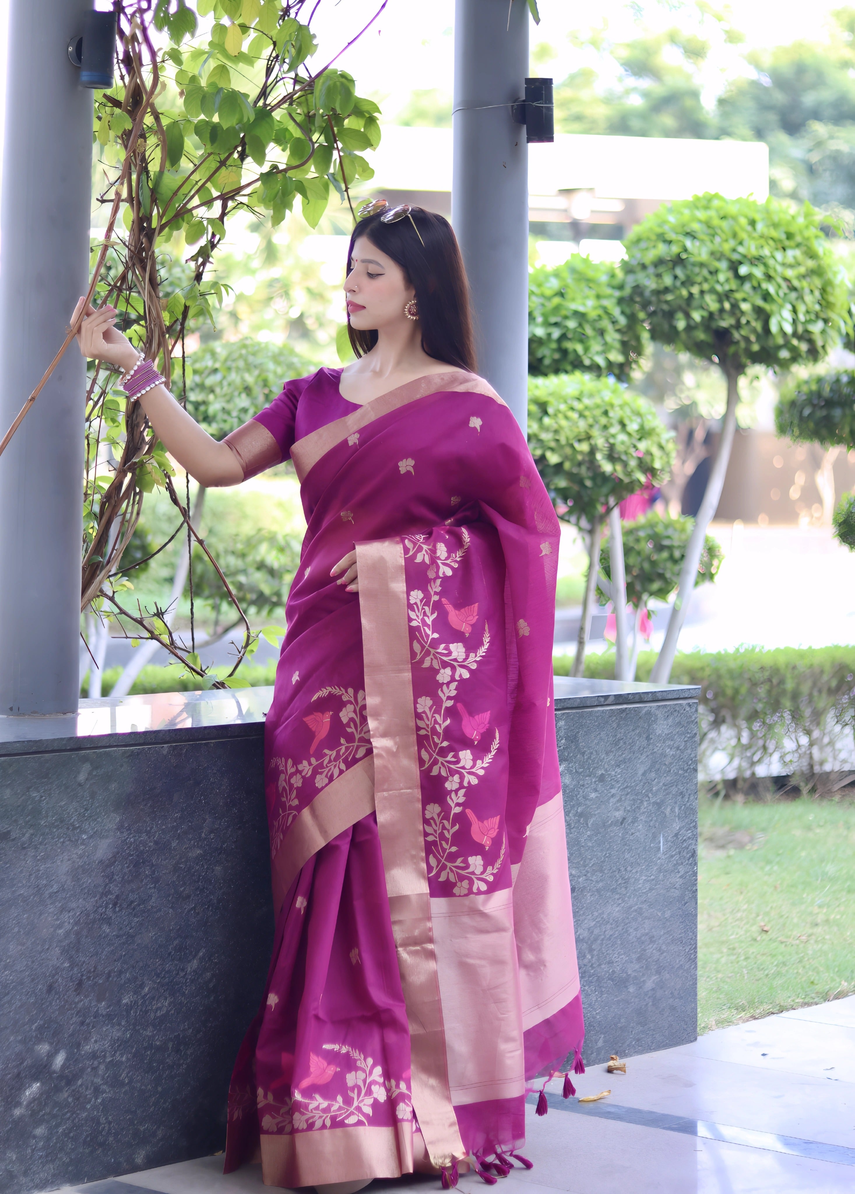 A woman models a vibrant Fuchsia Purple Linen Silk Saree with delicate gold Zari woven floral motifs. The saree has a wide metallic gold border. She wears a matching purple blouse and subtle gold jewelry, posing in a modern outdoor setting.