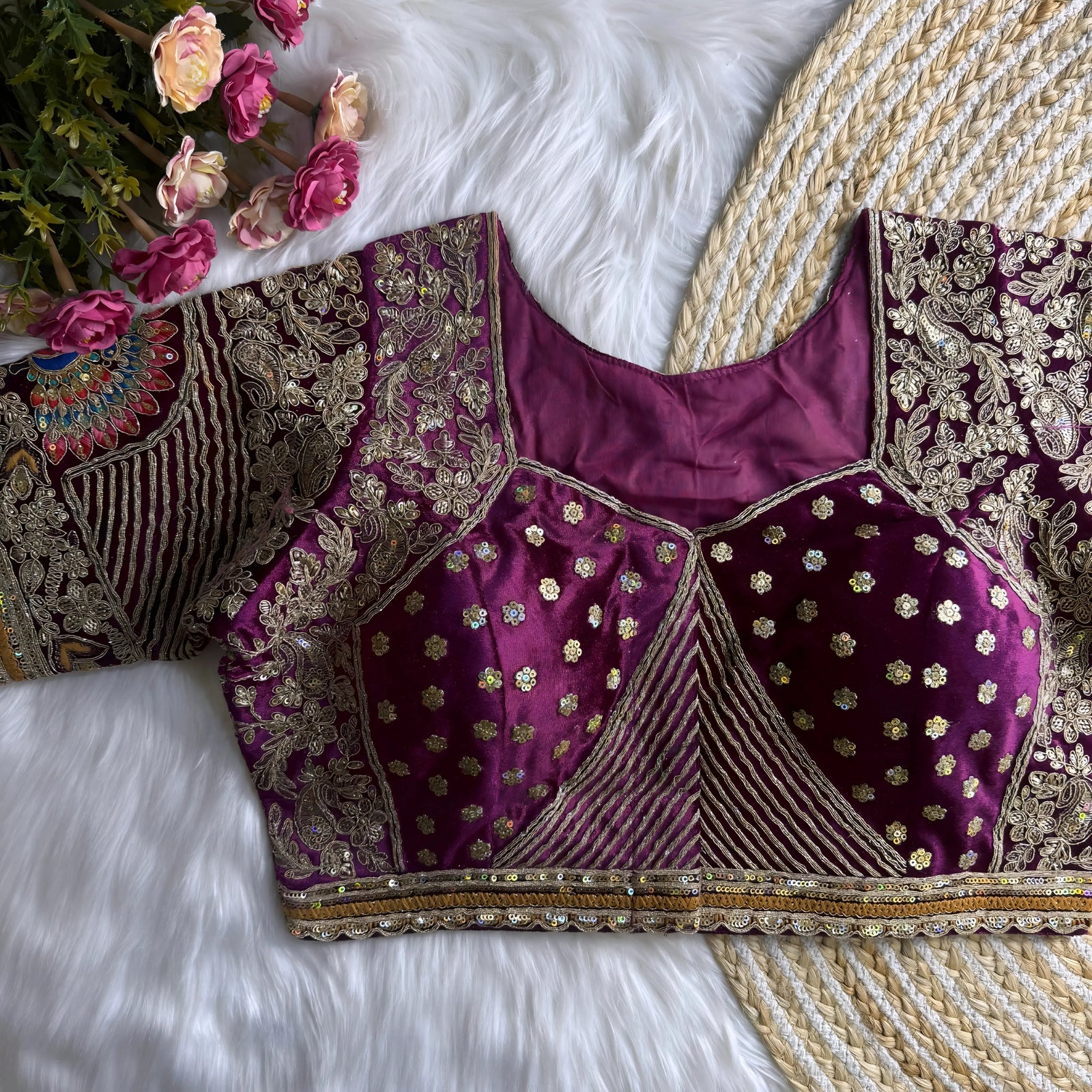 Traditional magenta silk blouse featuring intricate gold zardosi and thread embroidery with floral and peacock motifs, sequin embellishments, geometric panel designs, and detailed sleeves, displayed flat on a white fur surface with a braided mat and artificial pink flowers in the corner.