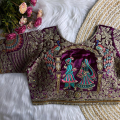 Traditional magenta silk blouse featuring intricate gold zardosi and thread embroidery with floral and peacock motifs, sequin embellishments, geometric panel designs, and detailed sleeves, displayed flat on a white fur surface with a braided mat and artificial pink flowers in the corner.