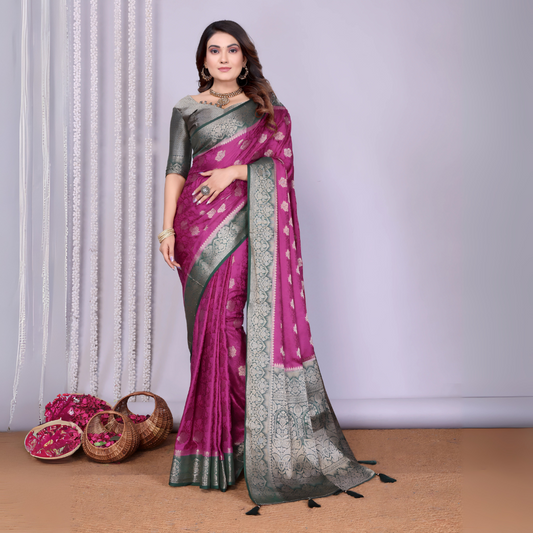 Woman wearing a magenta Banarasi silk saree with a silver and grey zari border, intricate woven motifs, and a matching grey blouse. She is adorned with traditional jewelry and poses gracefully in a modern studio setting featuring woven baskets filled with rose petals and white curtain drapes against a neutral backdrop.