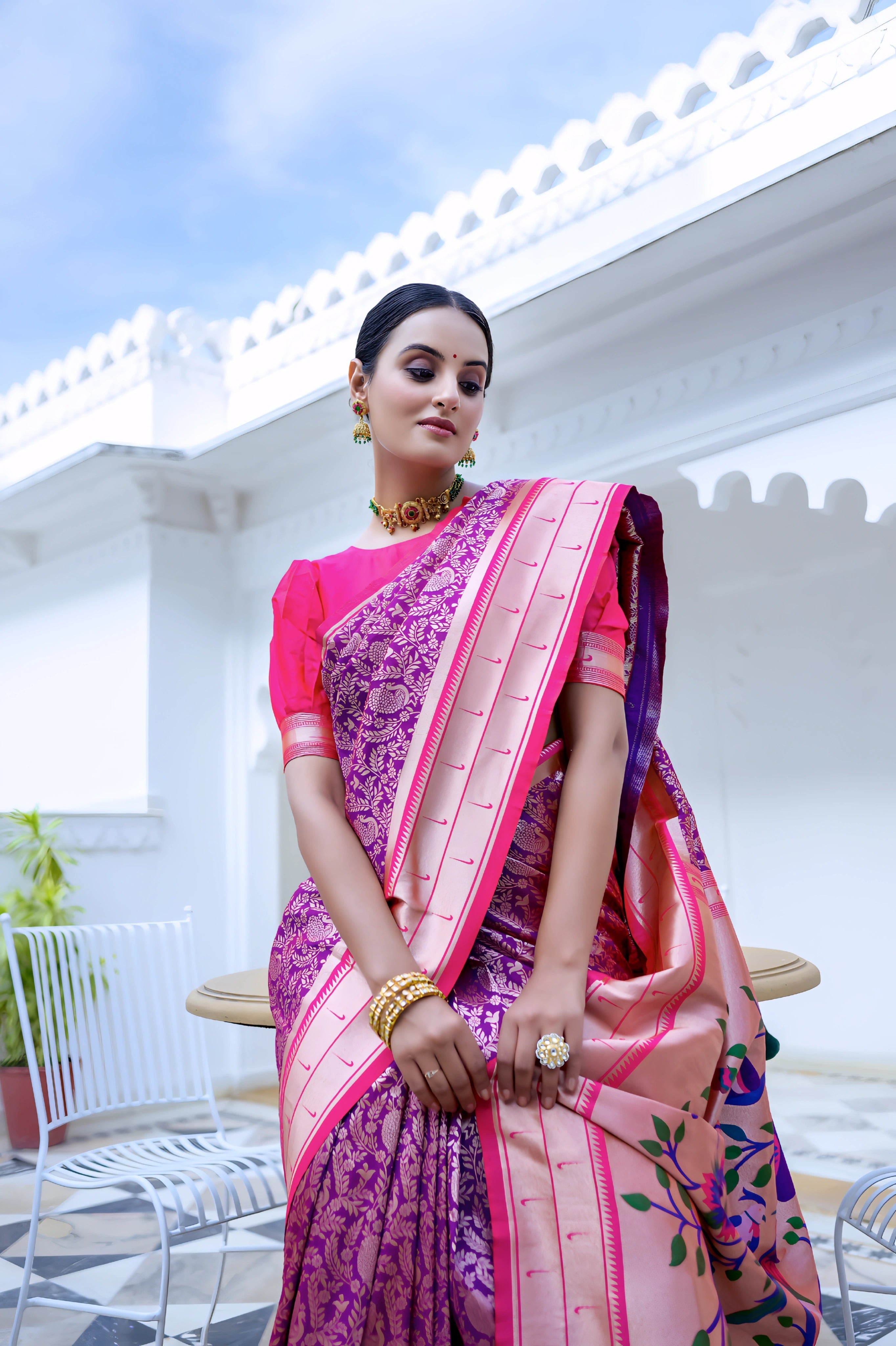 A woman models a vibrant Royal Purple Soft Silk Saree. The saree body features subtle woven patterns. It has a striking light pink contrast border with gold Zari. The pallu is a focal point, featuring rich, artistic Paithani-style print or weaving of colorful bird-on-branch motifs. She wears a fuchsia pink blouse and traditional gold jewelry, posing on an ornate white terrace.