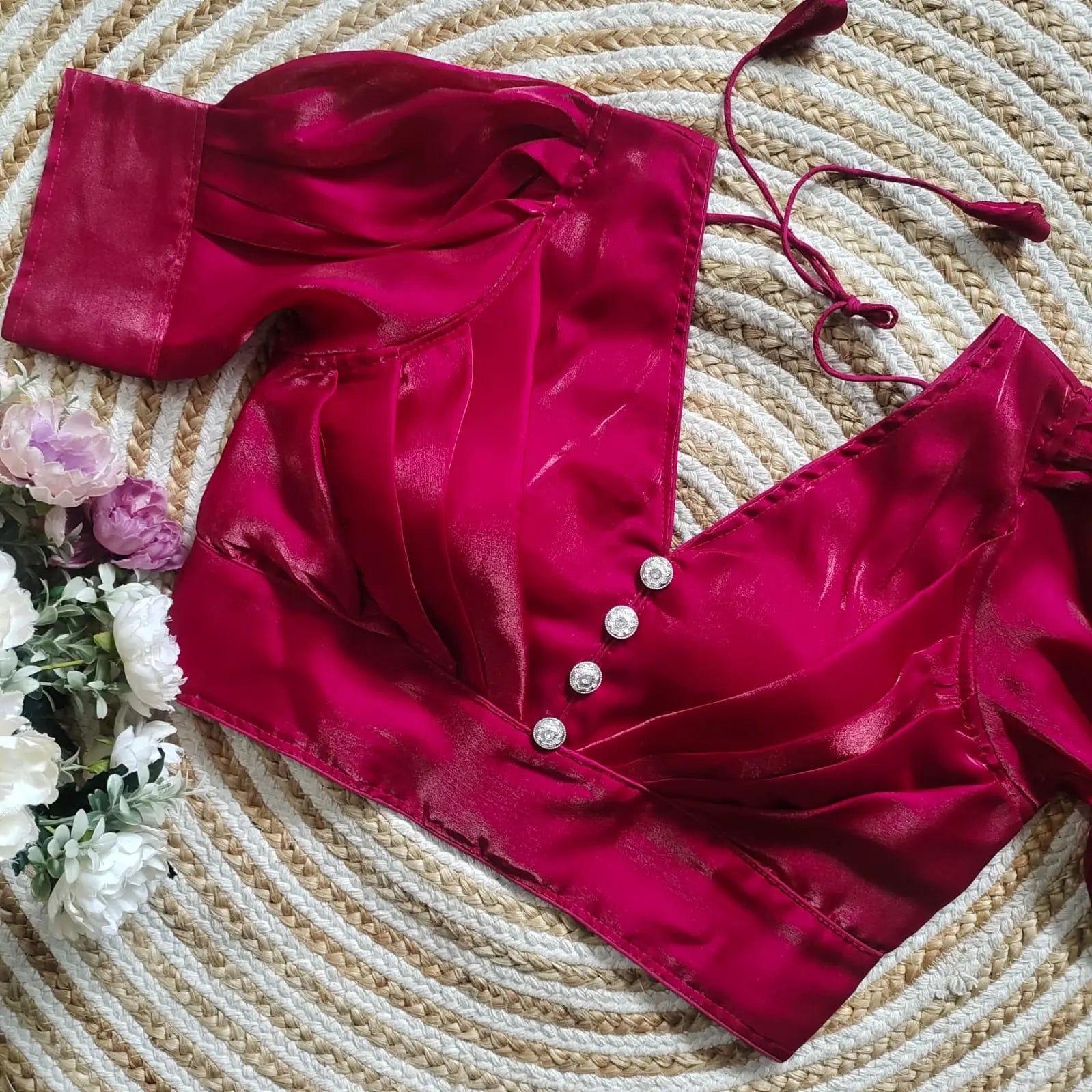Magenta satin blouse with pleated detailing and four decorative silver buttons on the front, featuring elbow-length sleeves and tie-up tassels, displayed on a circular woven mat with white and pink flowers on the side.
