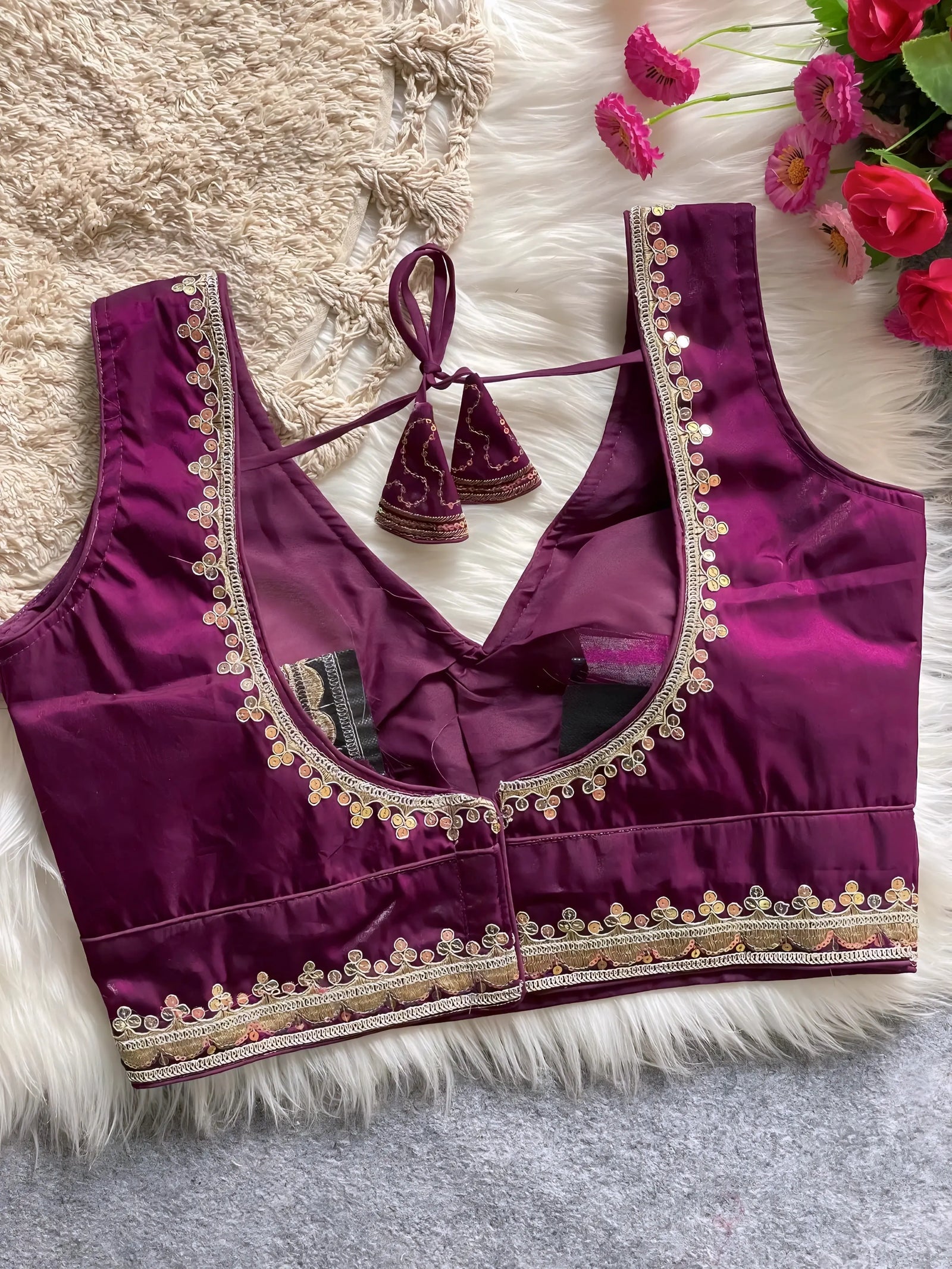 Traditional magenta silk blouse featuring intricate gold embroidery and beadwork with floral motifs along the neckline and hem, displayed flat on a soft fur and textured mat background with pink artificial flowers in the corner.