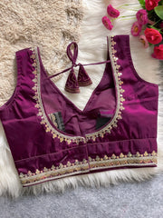 Traditional magenta silk blouse featuring intricate gold embroidery and beadwork with floral motifs along the neckline and hem, displayed flat on a soft fur and textured mat background with pink artificial flowers in the corner.
