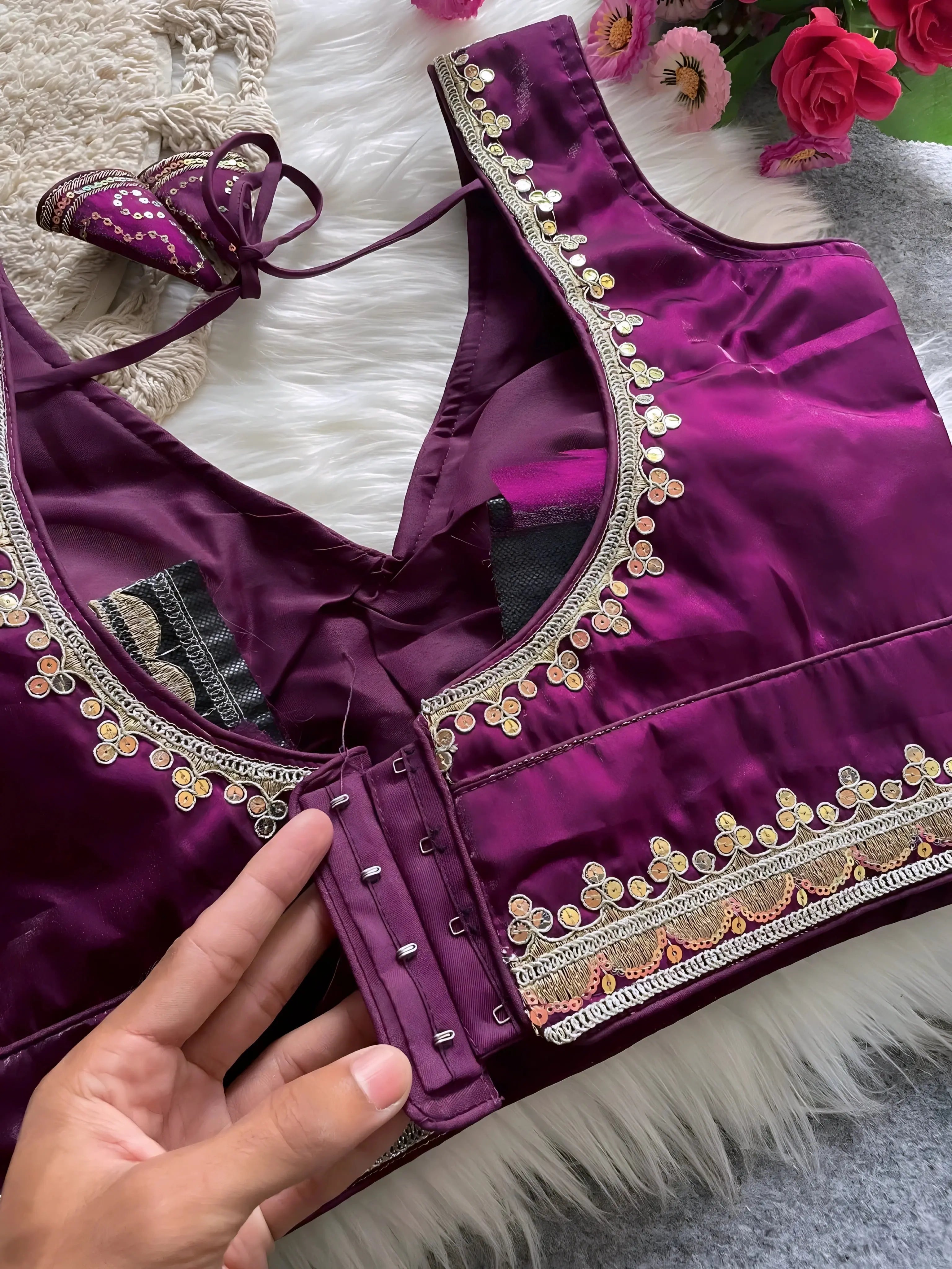 Traditional magenta silk blouse featuring intricate gold embroidery and beadwork with floral motifs along the neckline and hem, displayed flat on a soft fur and textured mat background with pink artificial flowers in the corner.