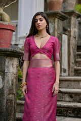 A woman models a stunning Rani Pink Soft Silk Saree. The saree features a wide, dual-toned metallic gold and pink Zari woven border and subtle metallic motifs on the body. She wears a matching pink short-sleeve blouse and a gold choker necklace, posing outdoors on an ornate stone staircase.