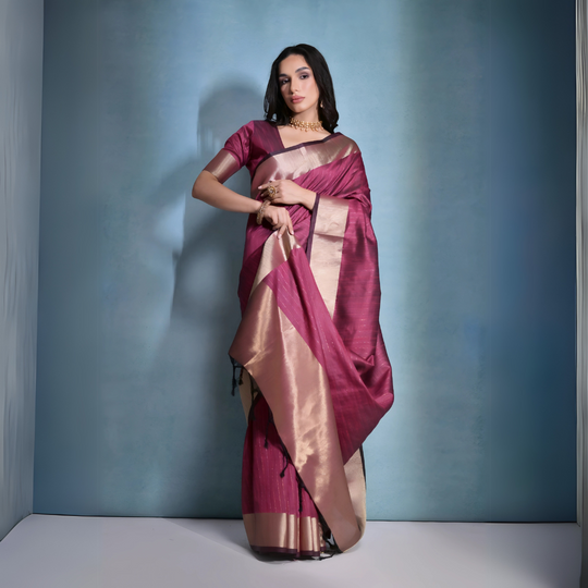 A woman models a striking Wine Red Banarasi Raw Silk Saree. The saree body features subtle woven stripes and is framed by an extremely wide, luxurious metallic gold Zari border. She wears a matching wine-colored blouse and traditional gold Kundan jewelry.