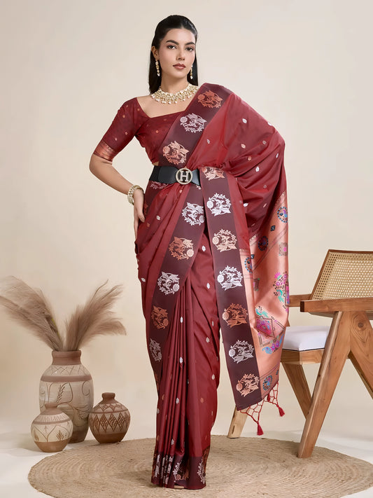A woman models a luxurious Dark Maroon Soft Silk Saree. The saree features scattered metallic motifs and a wide, contrasting peach/copper pallu panel richly woven with multi-colored traditional figurative and floral designs. She wears a matching brown blouse and heavy gold Kundan jewelry, posing indoors.