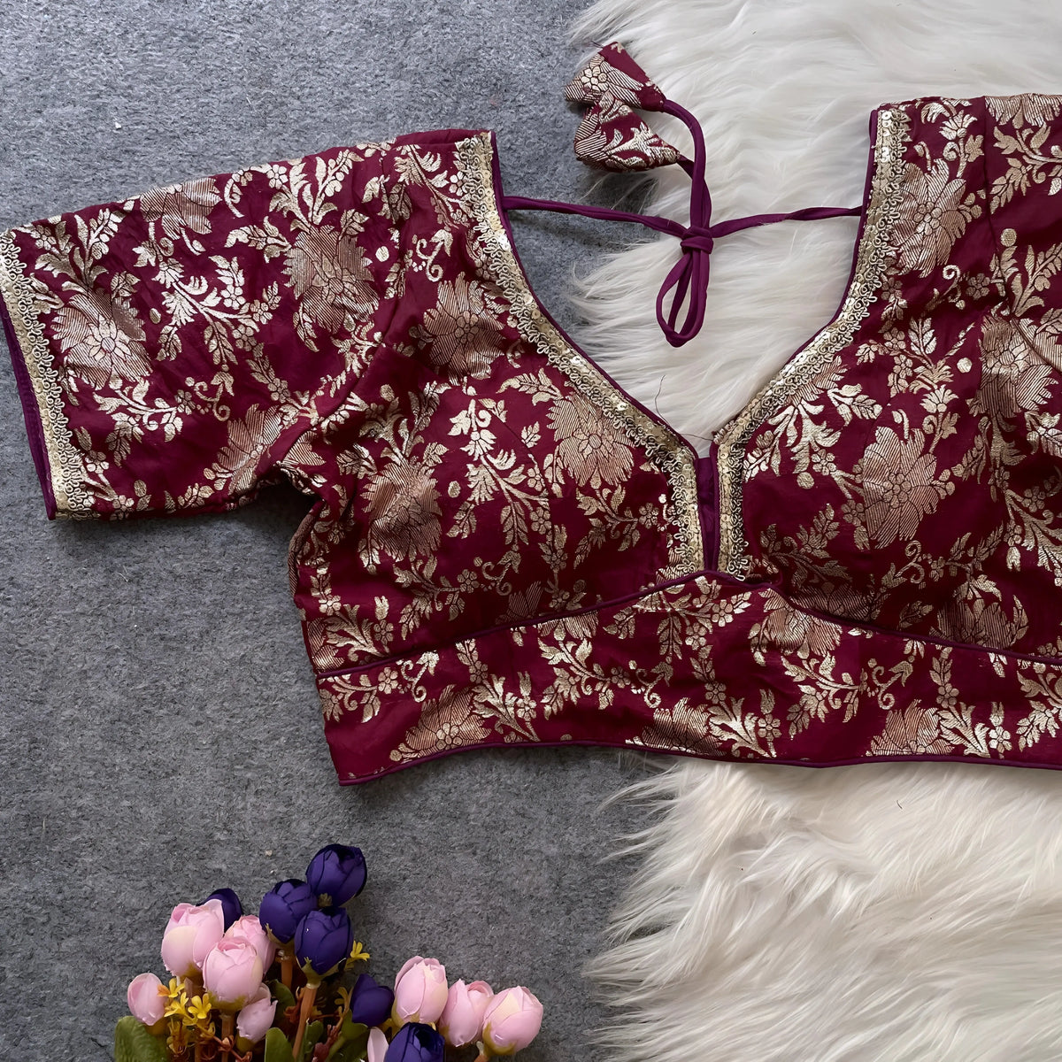 Maroon brocade blouse featuring intricate gold floral zari embroidery, decorative trim along the neckline, and short sleeves, displayed flat on a grey surface with a white faux fur accent and a bunch of artificial pink and purple flowers for an elegant, festive ethnic look.
