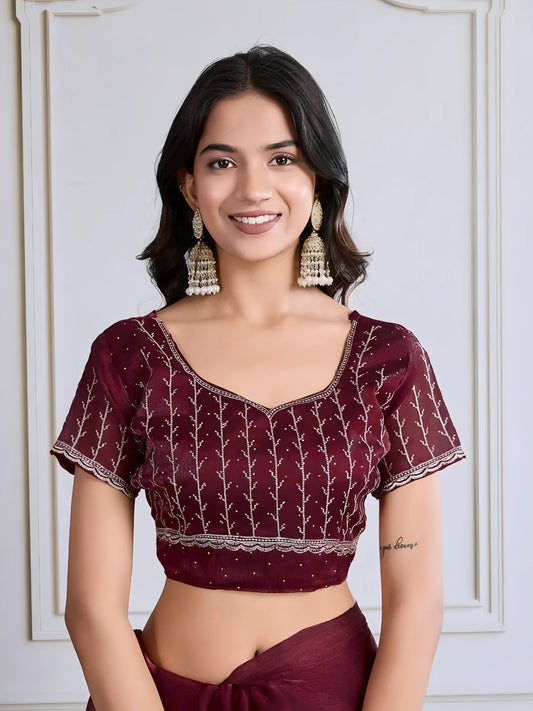 Woman wearing a maroon georgette saree adorned with delicate sequins and an ornate silver embroidered border, paired with a matching blouse and traditional jhumka earrings. She stands elegantly in a classic white paneled room with a vintage rotary telephone on a carved wooden side table, a modern wooden chair with a woven seat, and a patterned rug, creating a sophisticated and timeless ambiance.