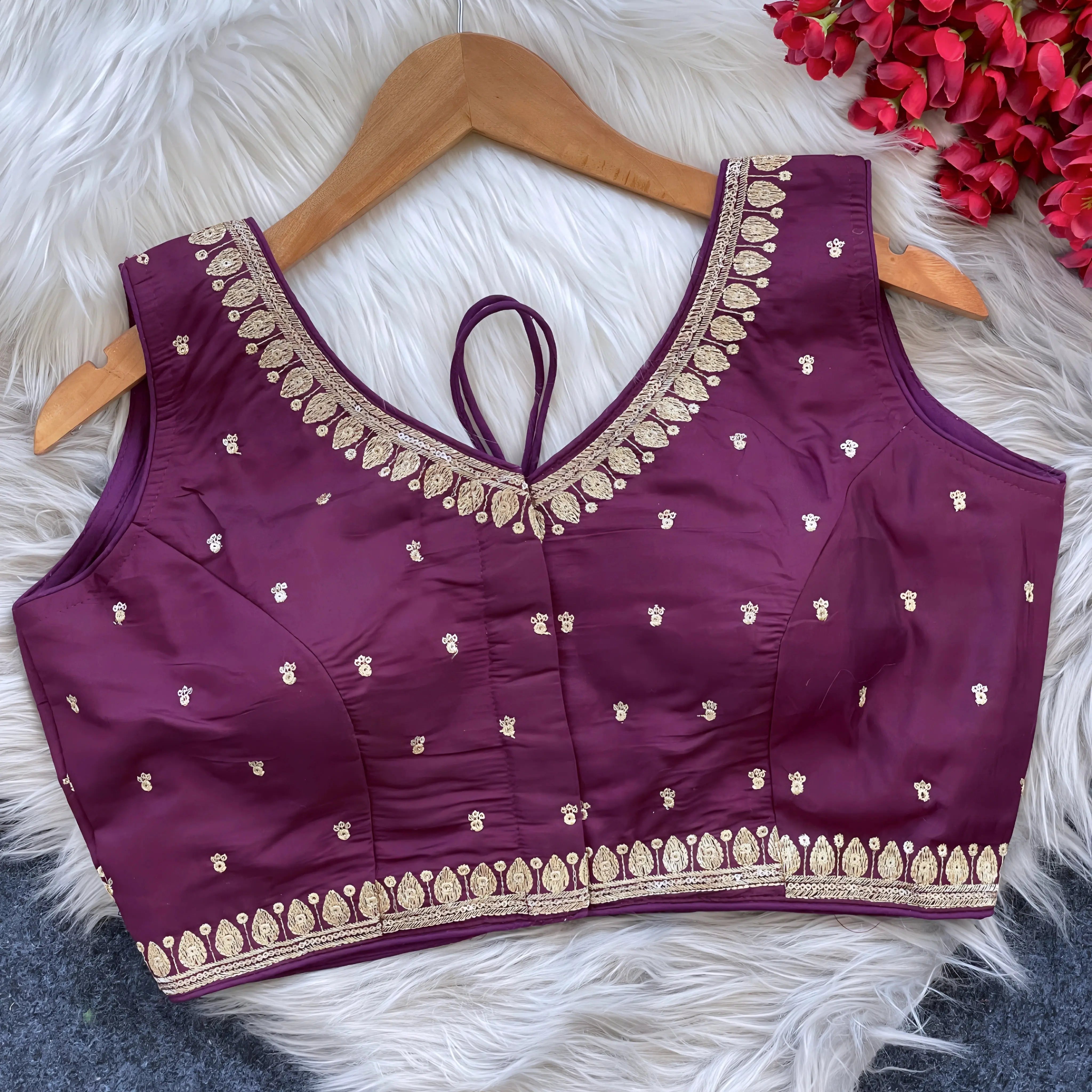 Maroon padded blouse with intricate gold embroidery along the neckline and hem, featuring small gold embroidered motifs, displayed on a wooden hanger over a white fur surface with red artificial flowers in the background.
