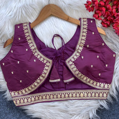 Maroon padded blouse with intricate gold embroidery along the neckline and hem, featuring small gold embroidered motifs, displayed on a wooden hanger over a white fur surface with red artificial flowers in the background.