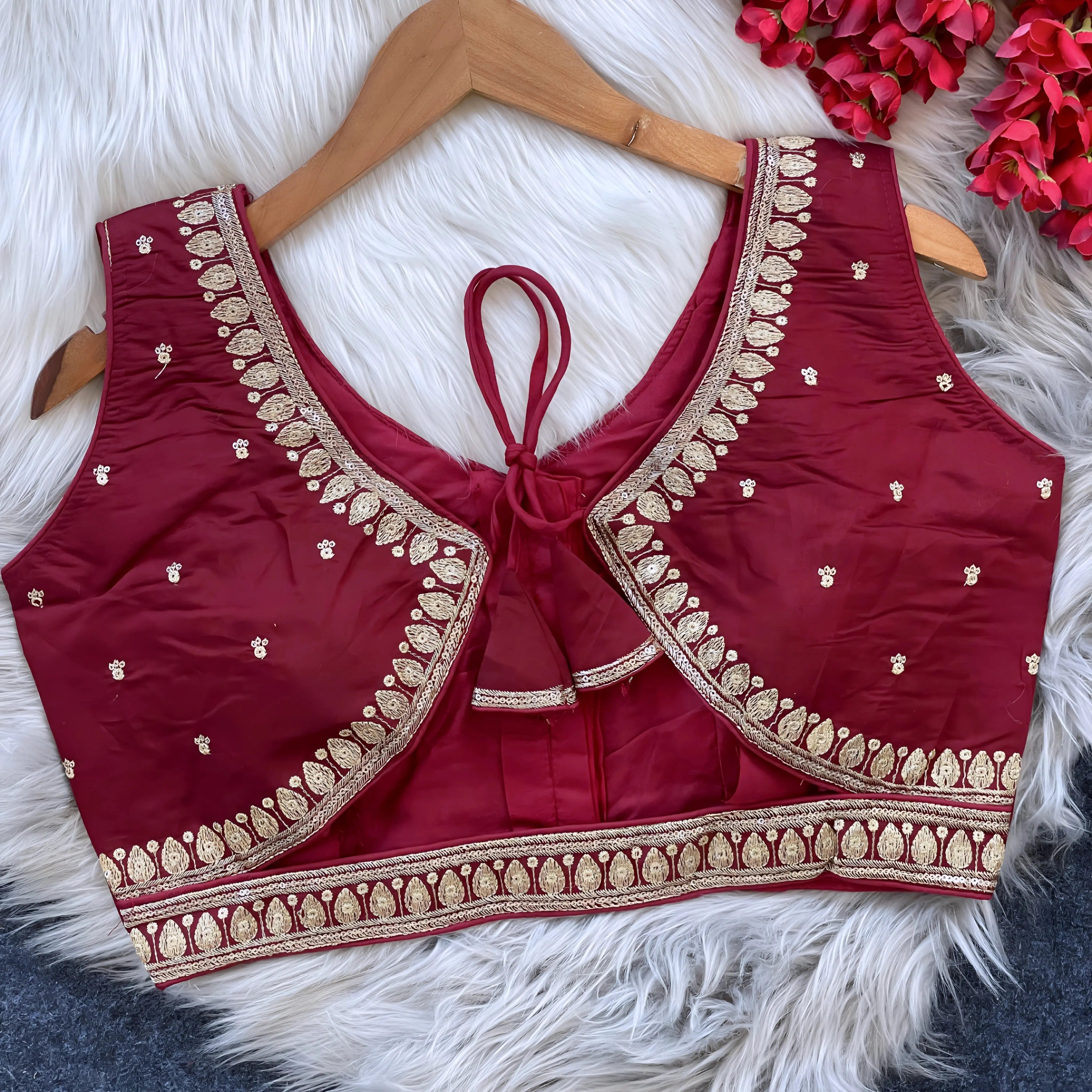 Maroon padded blouse with intricate gold embroidery along the neckline and hem, featuring small gold motifs, displayed on a wooden hanger over a white fur surface with red artificial flowers in the background.