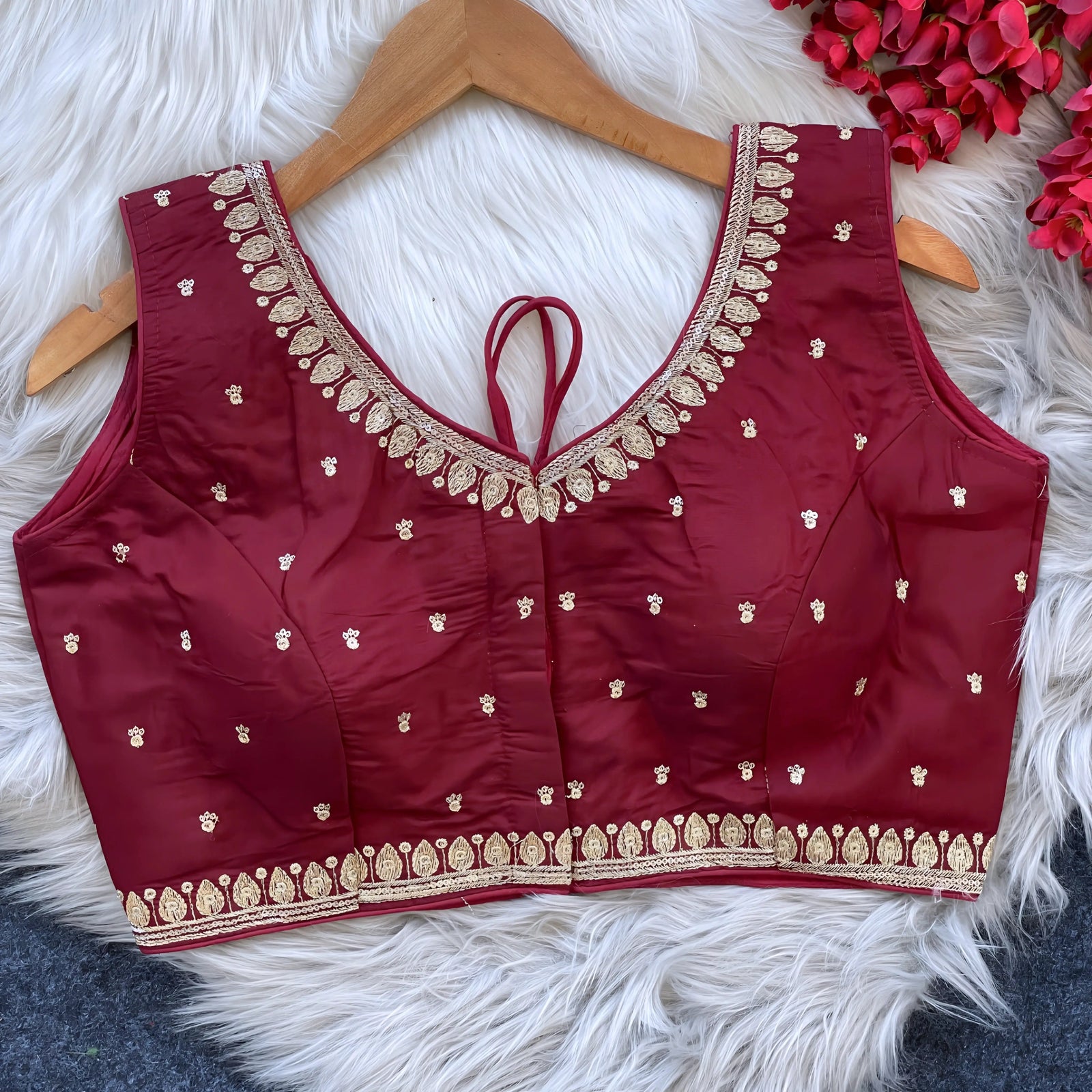 Maroon padded blouse with intricate gold embroidery along the neckline and hem, featuring small gold motifs, displayed on a wooden hanger over a white fur surface with red artificial flowers in the background.