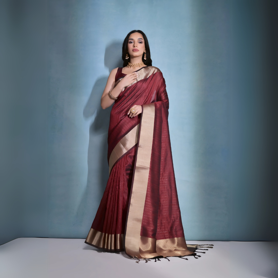 A woman models a striking Dark Maroon Banarasi Raw Silk Saree. The saree features a very wide, luxurious metallic gold Zari border and subtle woven stripes on the body. She wears a sleeveless maroon blouse and traditional gold Kundan jewelry, posing indoors.