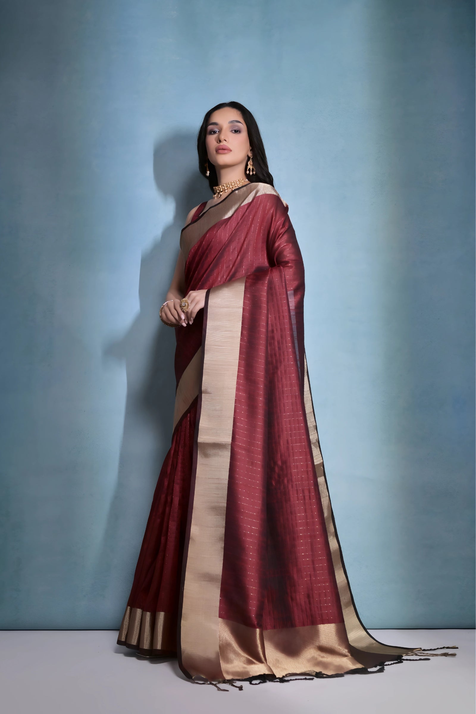 A woman models a striking Dark Maroon Banarasi Raw Silk Saree. The saree features a very wide, luxurious metallic gold Zari border and subtle woven stripes on the body. She wears a sleeveless maroon blouse and traditional gold Kundan jewelry, posing indoors.