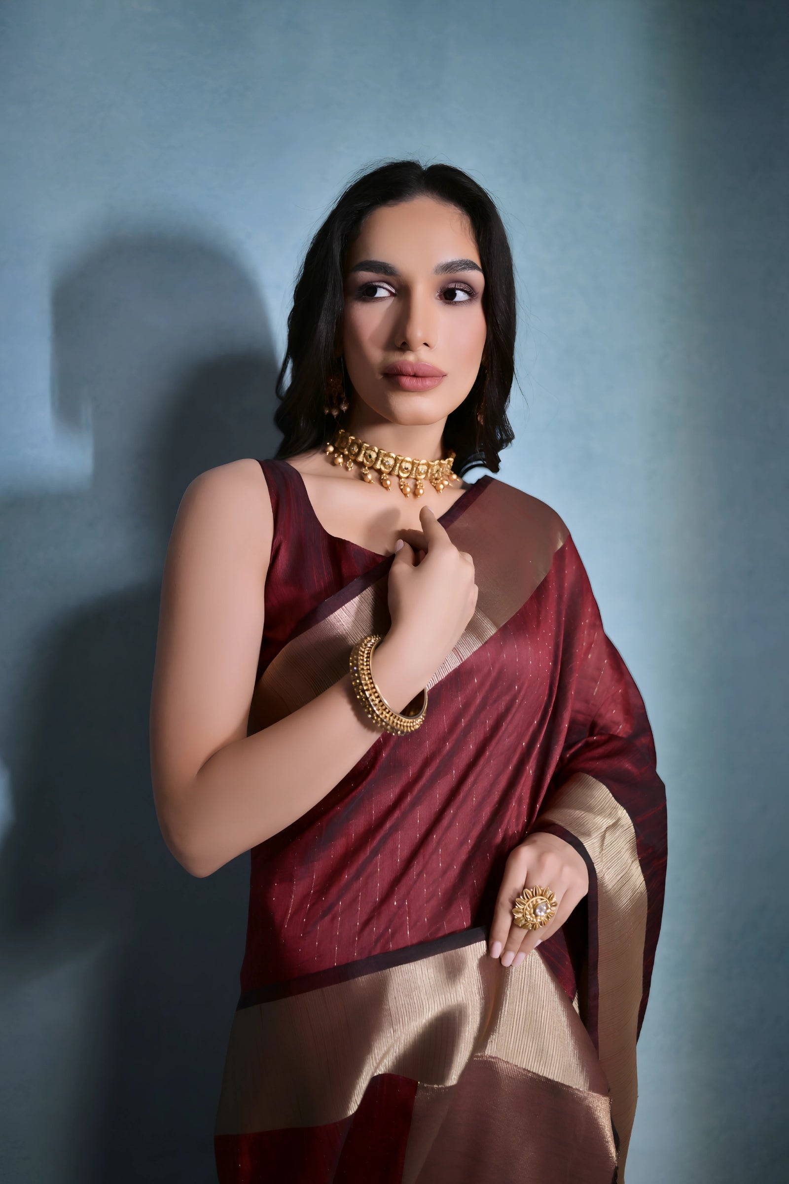 A woman models a striking Dark Maroon Banarasi Raw Silk Saree. The saree features a very wide, luxurious metallic gold Zari border and subtle woven stripes on the body. She wears a sleeveless maroon blouse and traditional gold Kundan jewelry, posing indoors.