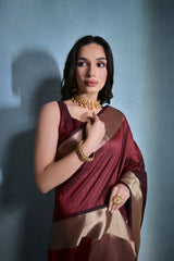 A woman models a striking Dark Maroon Banarasi Raw Silk Saree. The saree features a very wide, luxurious metallic gold Zari border and subtle woven stripes on the body. She wears a sleeveless maroon blouse and traditional gold Kundan jewelry, posing indoors.
