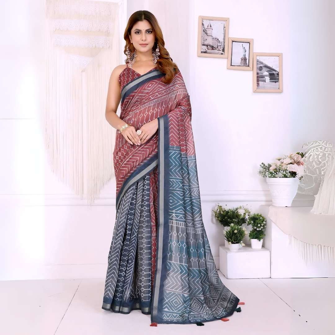 Woman wearing a maroon and grey cotton saree featuring bold geometric and tribal-inspired patterns on the pallu and pleats, paired with a sleeveless blouse and statement silver earrings. She poses elegantly in a contemporary studio setting with white walls, hanging macramé decor, floral arrangements, and framed minimalist artwork, showcasing the saree’s intricate design and graceful drape.