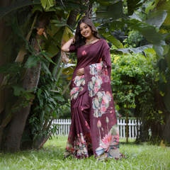 A woman models a stunning Deep Wine Soft Tussar Silk Saree. The saree features a large-scale digital print of colorful floral and lotus motifs in pink, white, and green on the pallu and lower drape. She wears a matching wine short-sleeve blouse and a gold choker necklace, posing outdoors in a bright garden setting.