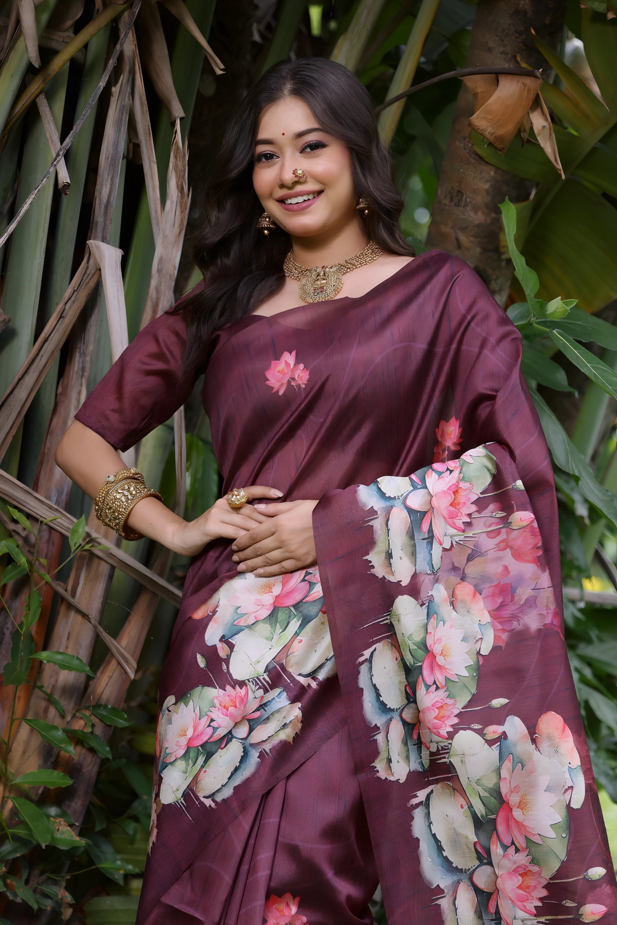 A woman models a stunning Deep Wine Soft Tussar Silk Saree. The saree features a large-scale digital print of colorful floral and lotus motifs in pink, white, and green on the pallu and lower drape. She wears a matching wine short-sleeve blouse and a gold choker necklace, posing outdoors in a bright garden setting.