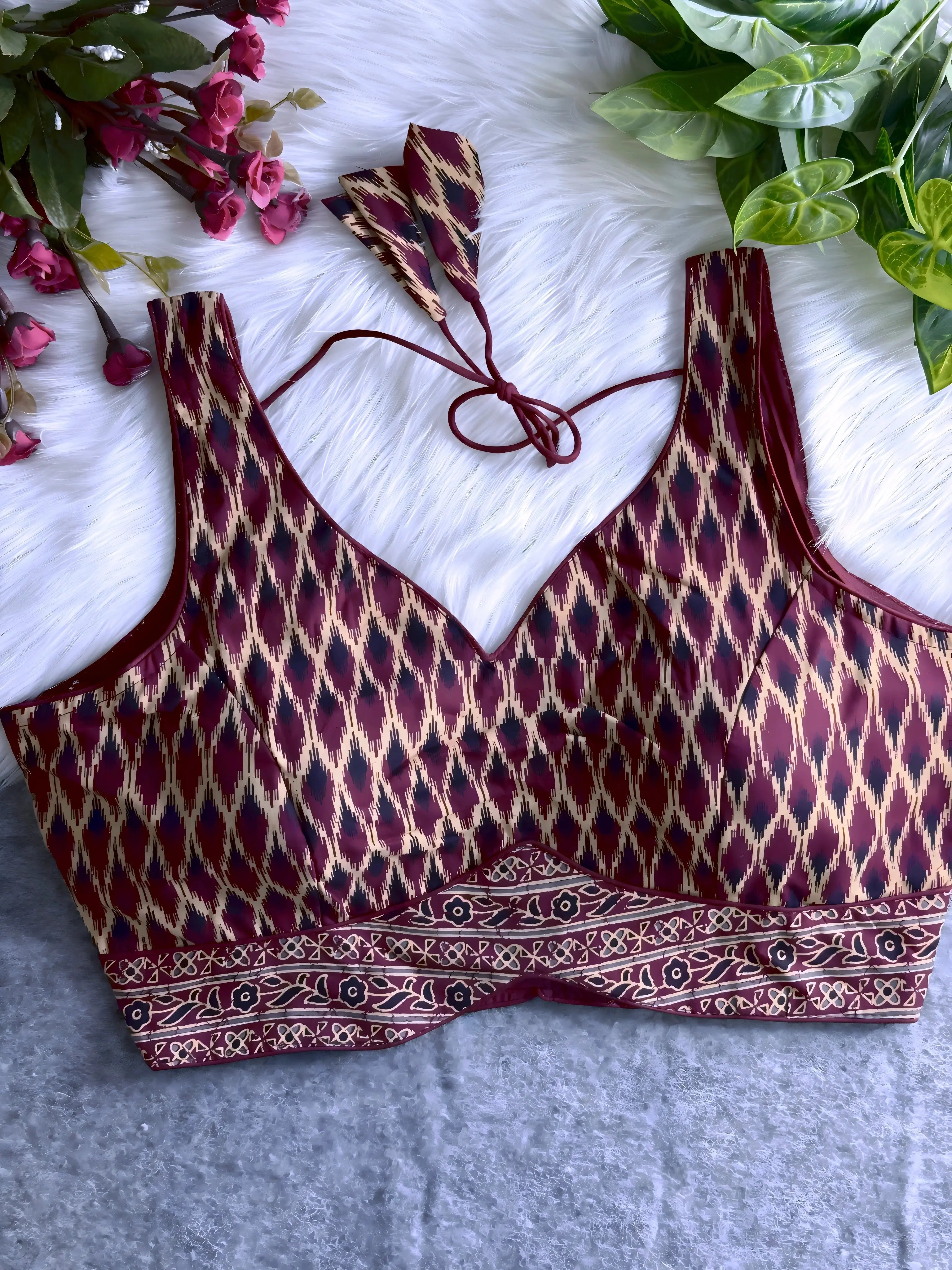 Maroon silk blouse featuring traditional Ikat-style geometric print in cream and black, wide shoulder straps, structured cups, and a patterned border at the hem. The design includes a V-neck and matching tasseled tie-back detail, displayed flat on a soft white fur background with artificial pink and green foliage arranged at the corners.
