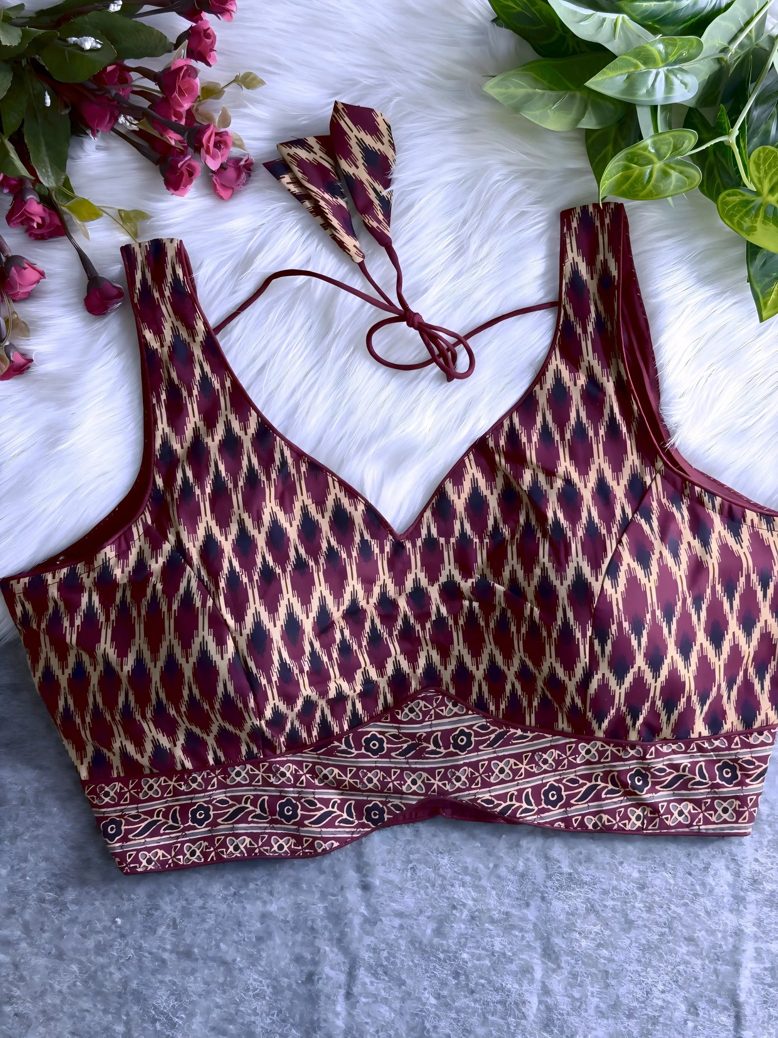 Maroon silk blouse featuring traditional Ikat-style geometric print in cream and black, wide shoulder straps, structured cups, and a patterned border at the hem. The design includes a V-neck and matching tasseled tie-back detail, displayed flat on a soft white fur background with artificial pink and green foliage arranged at the corners.