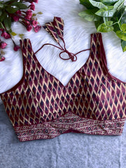 Maroon silk blouse featuring traditional Ikat-style geometric print in cream and black, wide shoulder straps, structured cups, and a patterned border at the hem. The design includes a V-neck and matching tasseled tie-back detail, displayed flat on a soft white fur background with artificial pink and green foliage arranged at the corners.